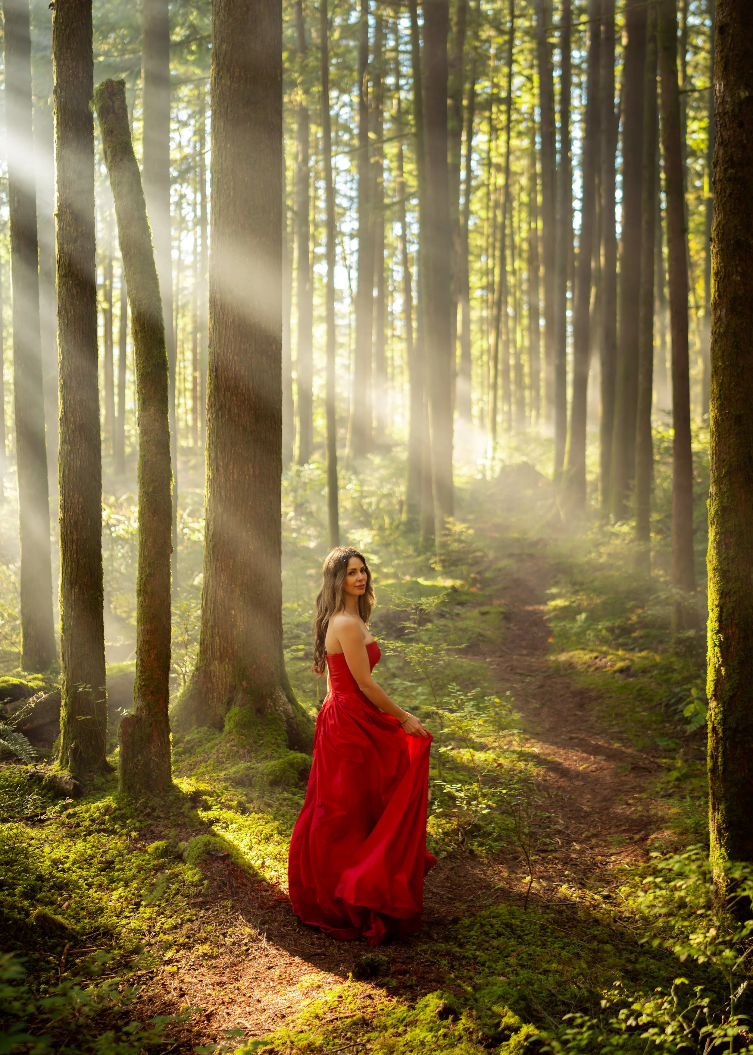 A woman in a flowing red dress standing on a dirt trail in a sunlit forest with tall trees and green moss.