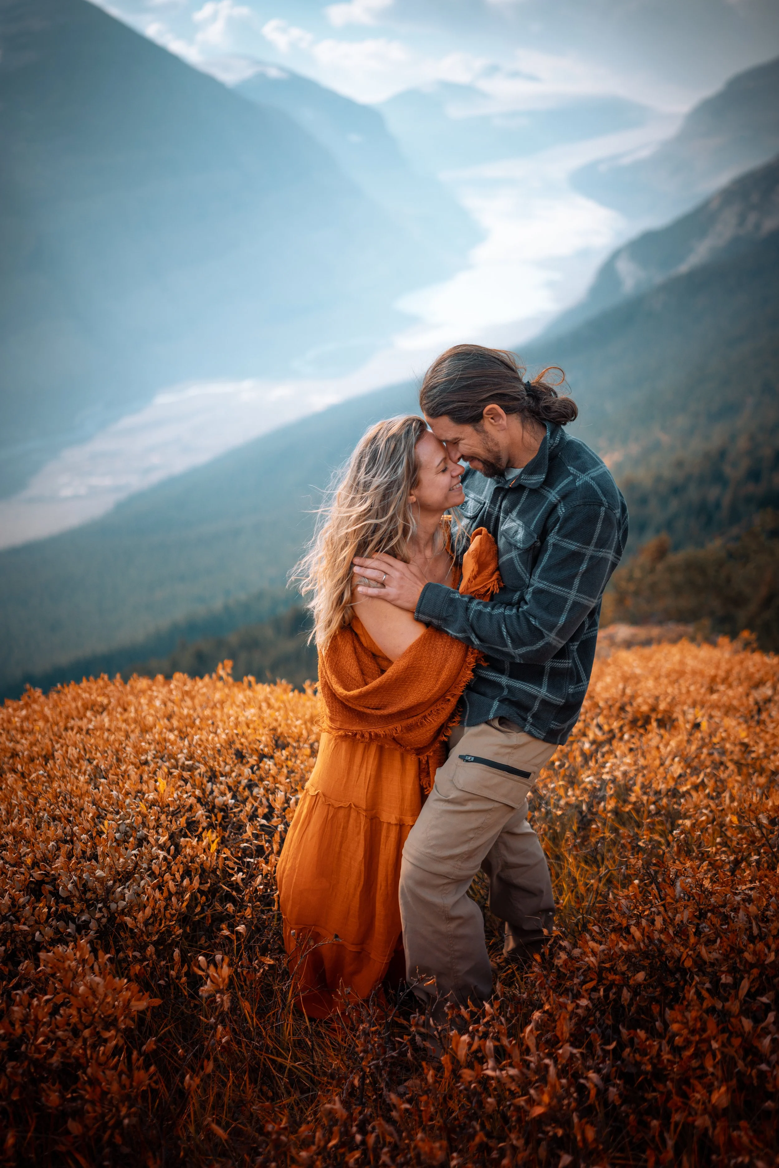A couple embracing on a mountain overlook surrounded by orange foliage with a river and mountain range in the background.