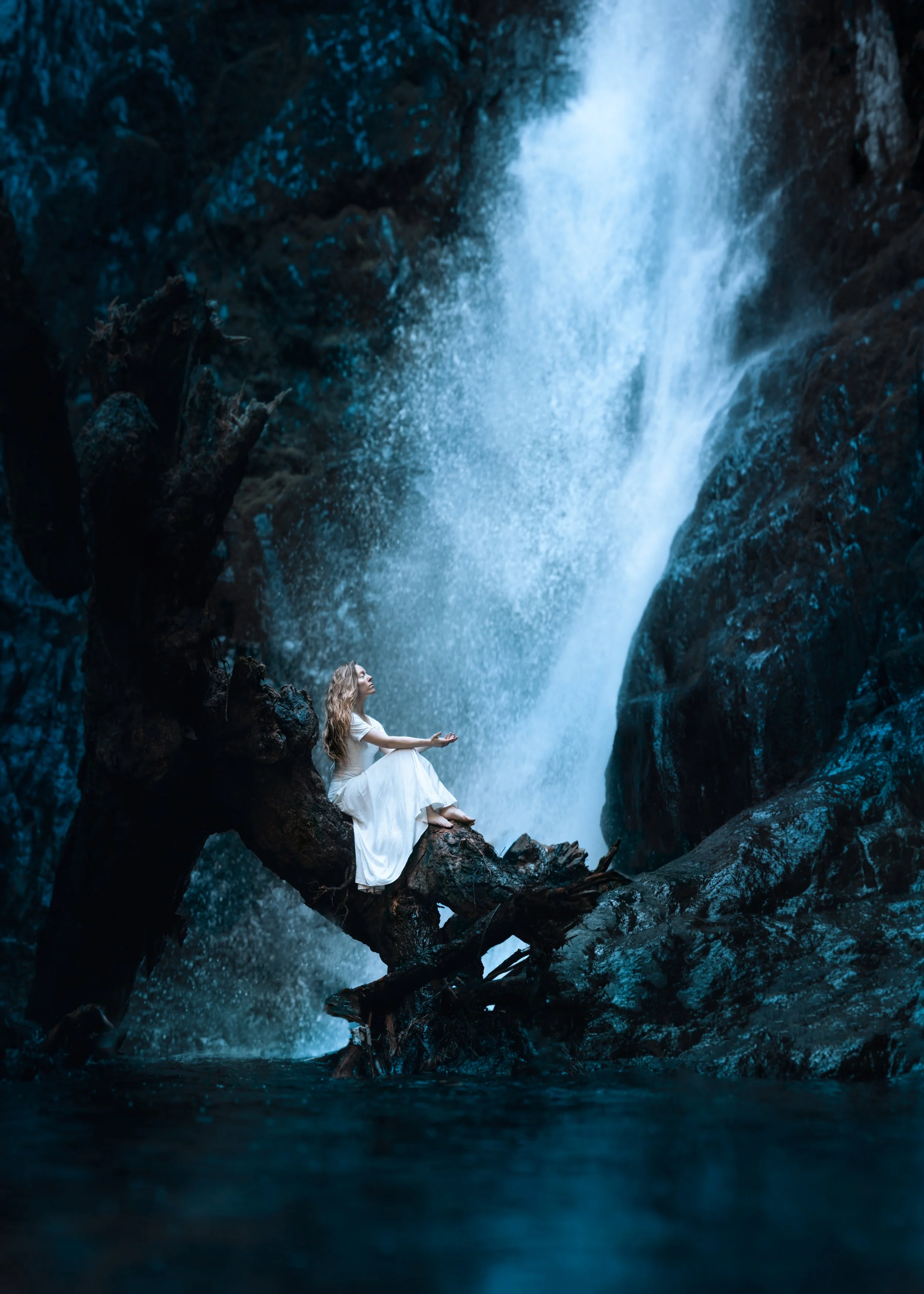 A woman in a white dress sitting on a large tree branch by a waterfall in a dark, rocky environment.