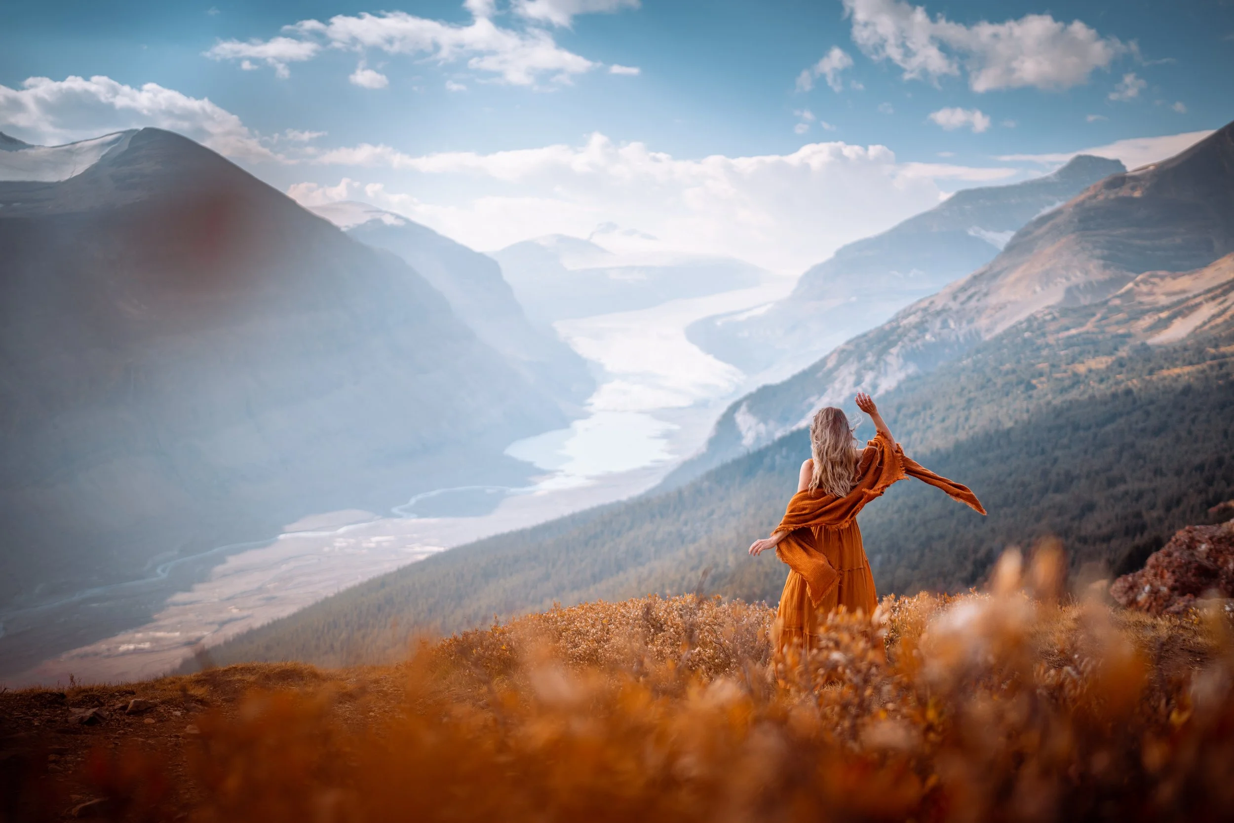 A woman in a flowing orange dress standing on a hillside, waving towards a scenic mountain valley with a river, surrounded by mountains and a partly cloudy sky.
