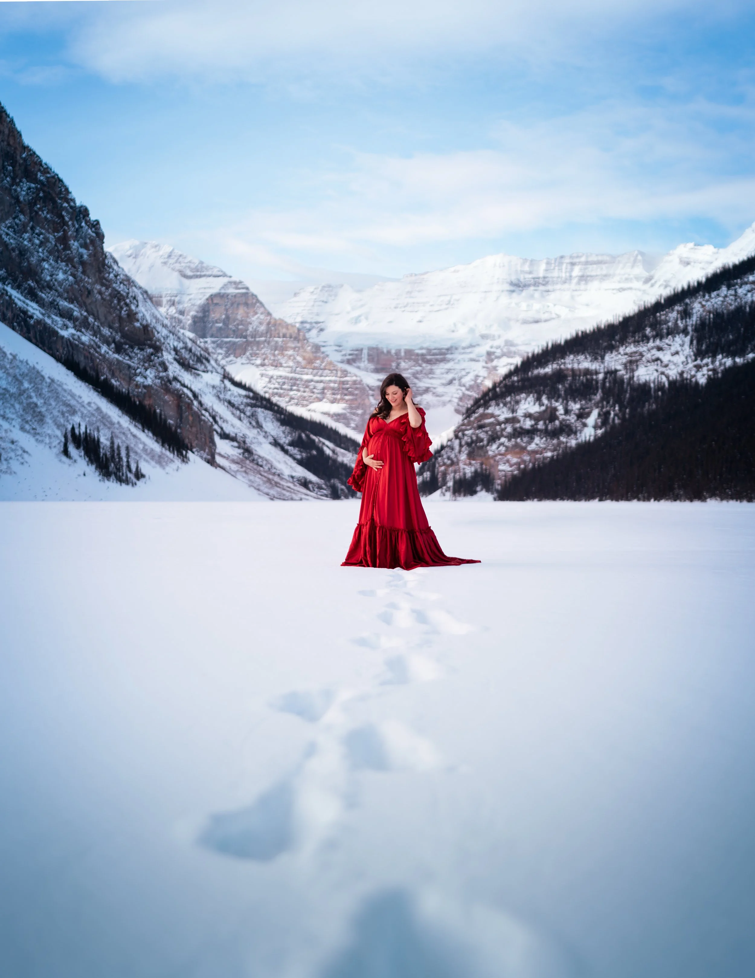 A pregnant woman in a long red dress standing in a snowy mountain landscape with snow-covered peaks and a partly cloudy sky in the background.