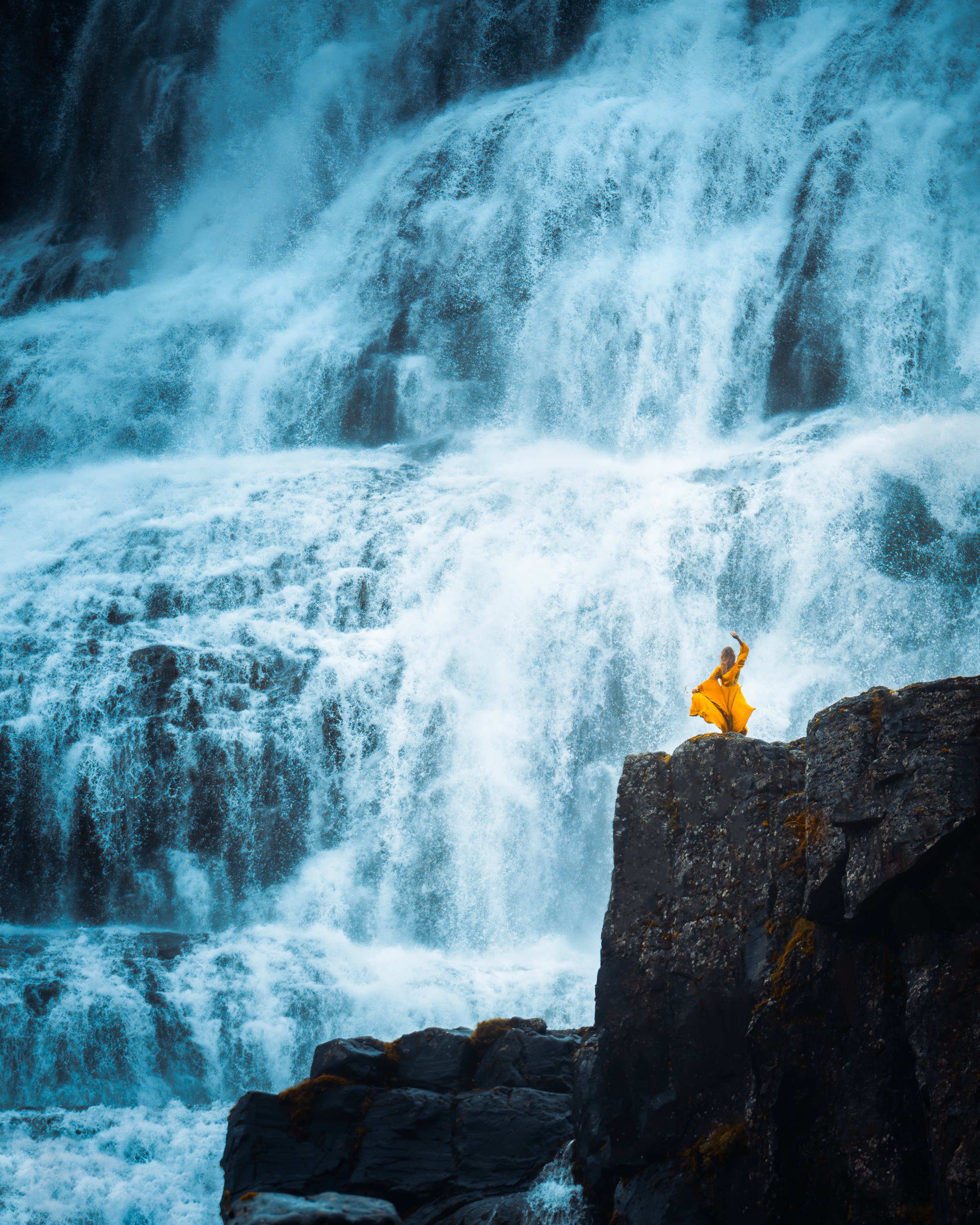 A person in a yellow raincoat standing on a rocky ledge near a large waterfall with cascading water.