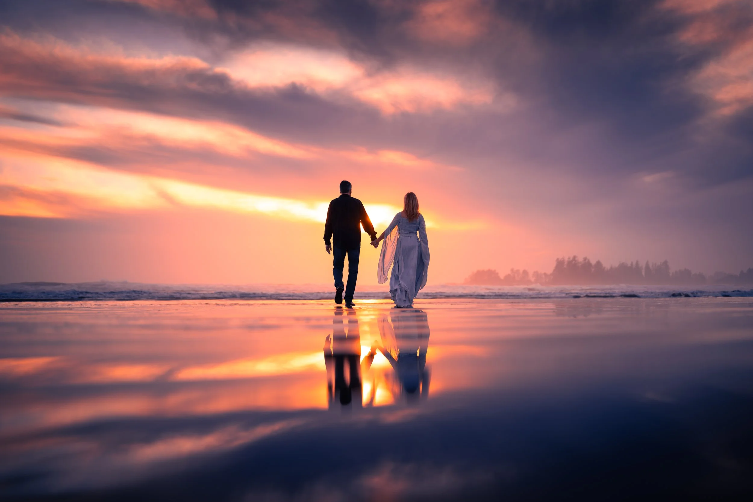 A couple holding hands walking on the beach during sunset with their reflection visible on the wet sand.