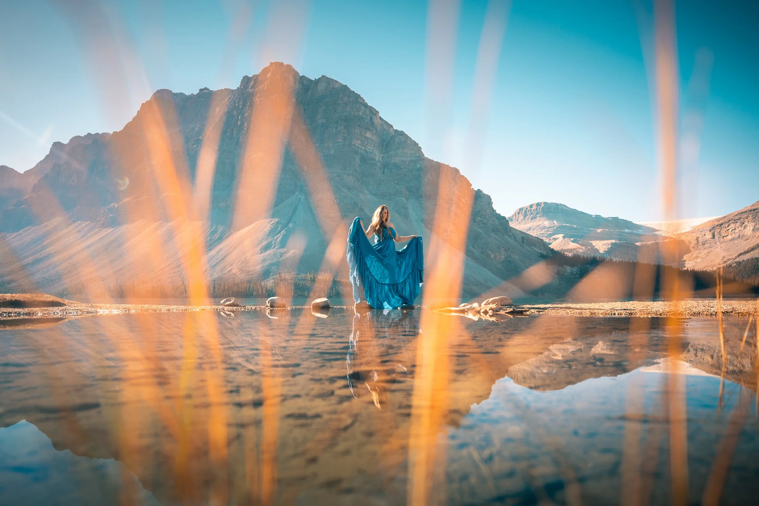 A woman in a blue dress standing in a shallow lake with mountains in the background, seen through tall grass or reeds.