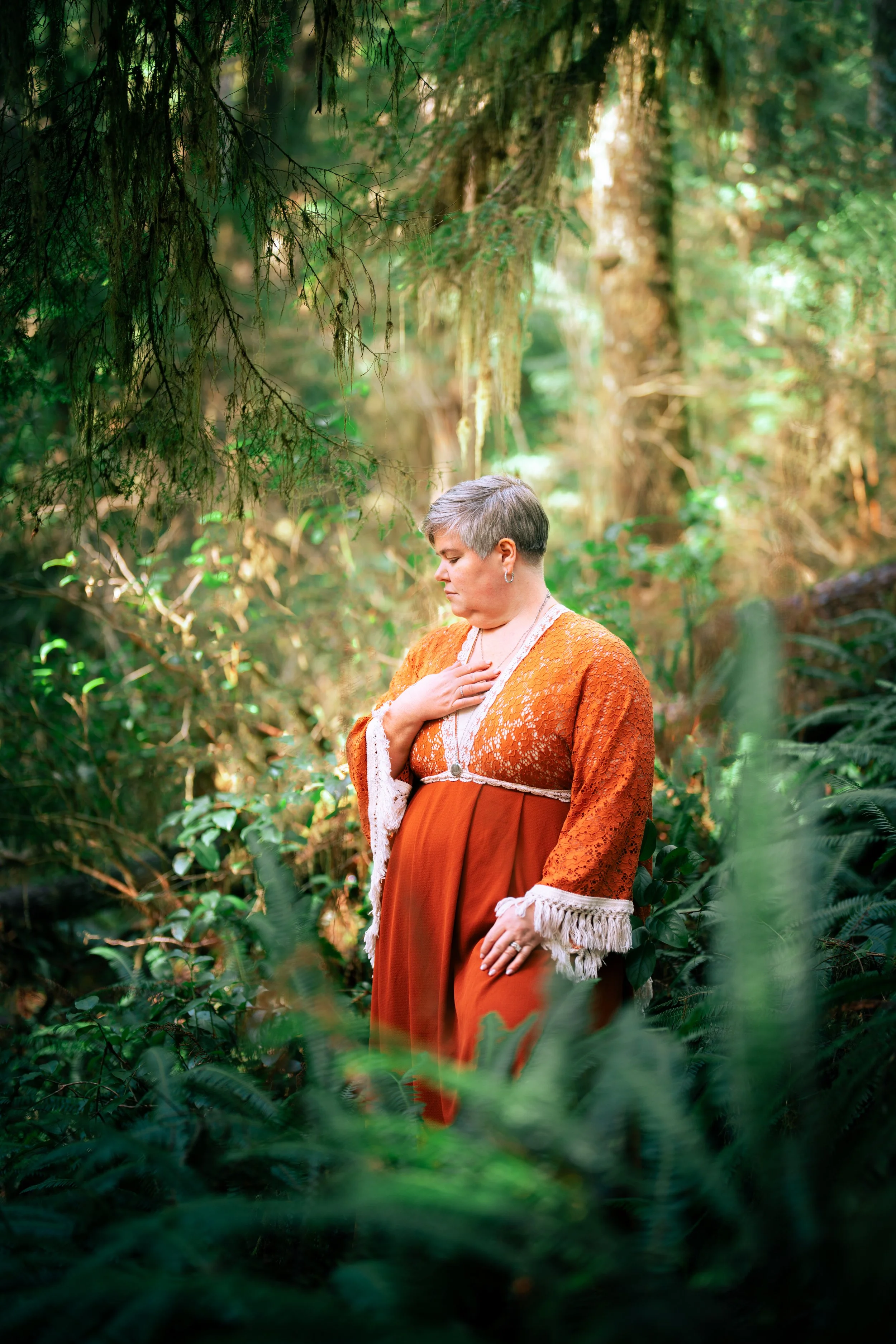 A woman with short gray hair wearing an orange and lace dress standing in a lush green forest, with her hand on her chest and her eyes closed.