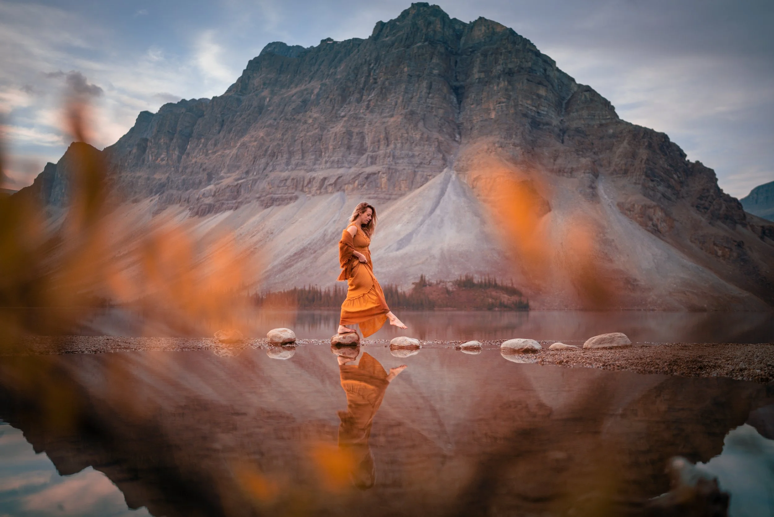 A woman in an orange dress walking barefoot across rocks in a calm lake with a mountain in the background during sunset or sunrise.