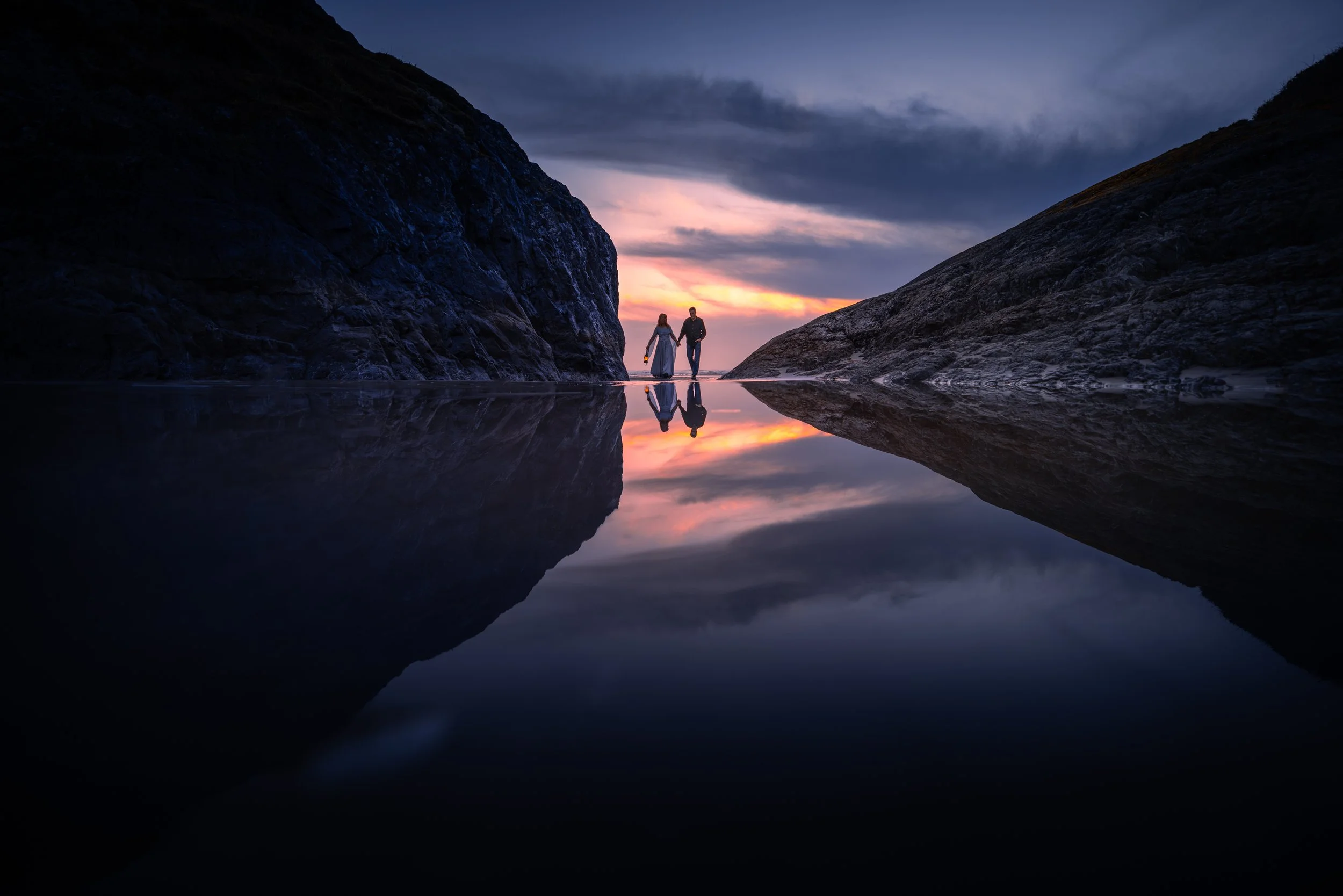 A couple walking on the beach at sunset, with large rocks on either side, and the water reflecting the sky and the couple.