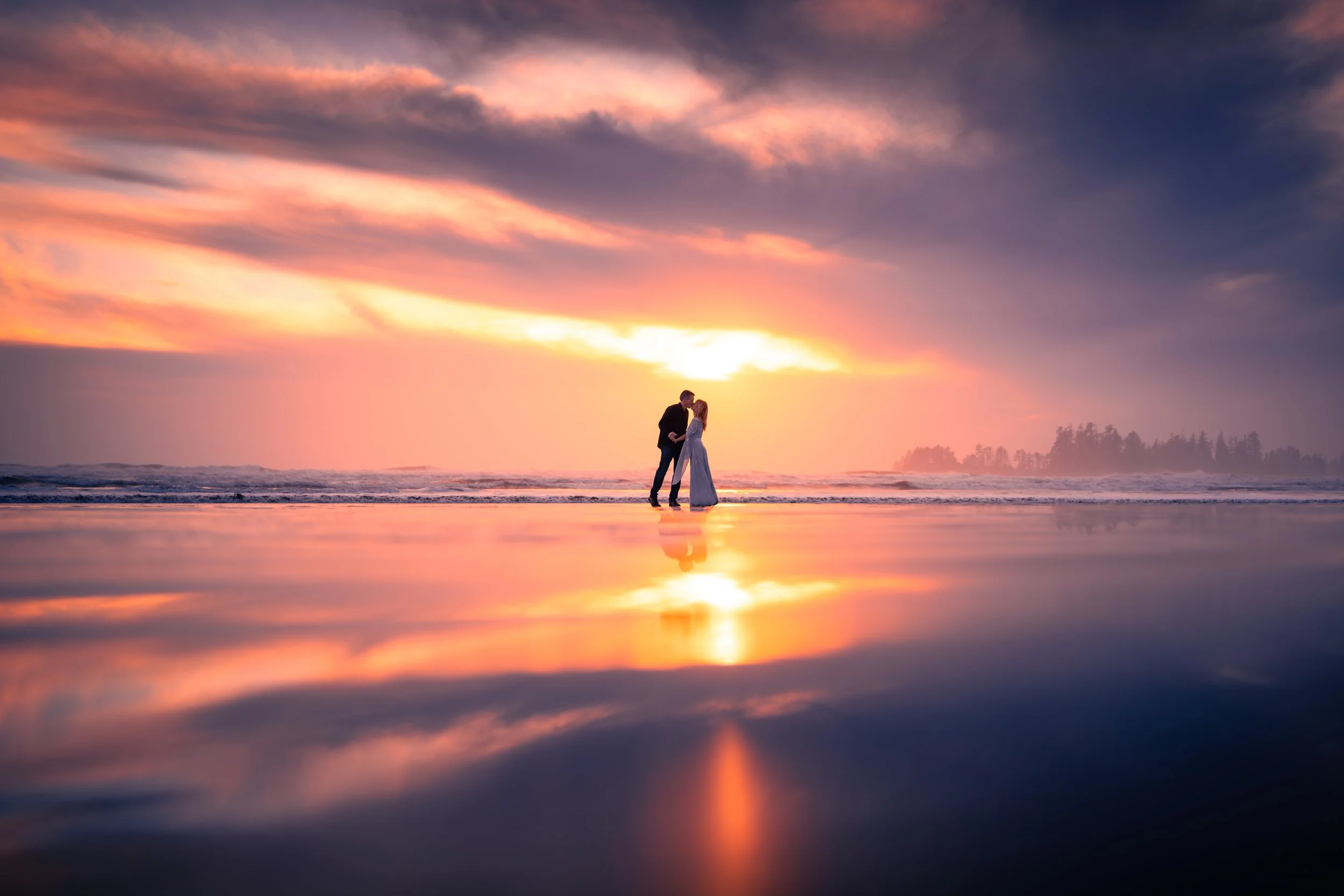 A couple standing on the beach at sunset, kissing, with the sky filled with pink and purple clouds, and their reflection visible on the wet sand.