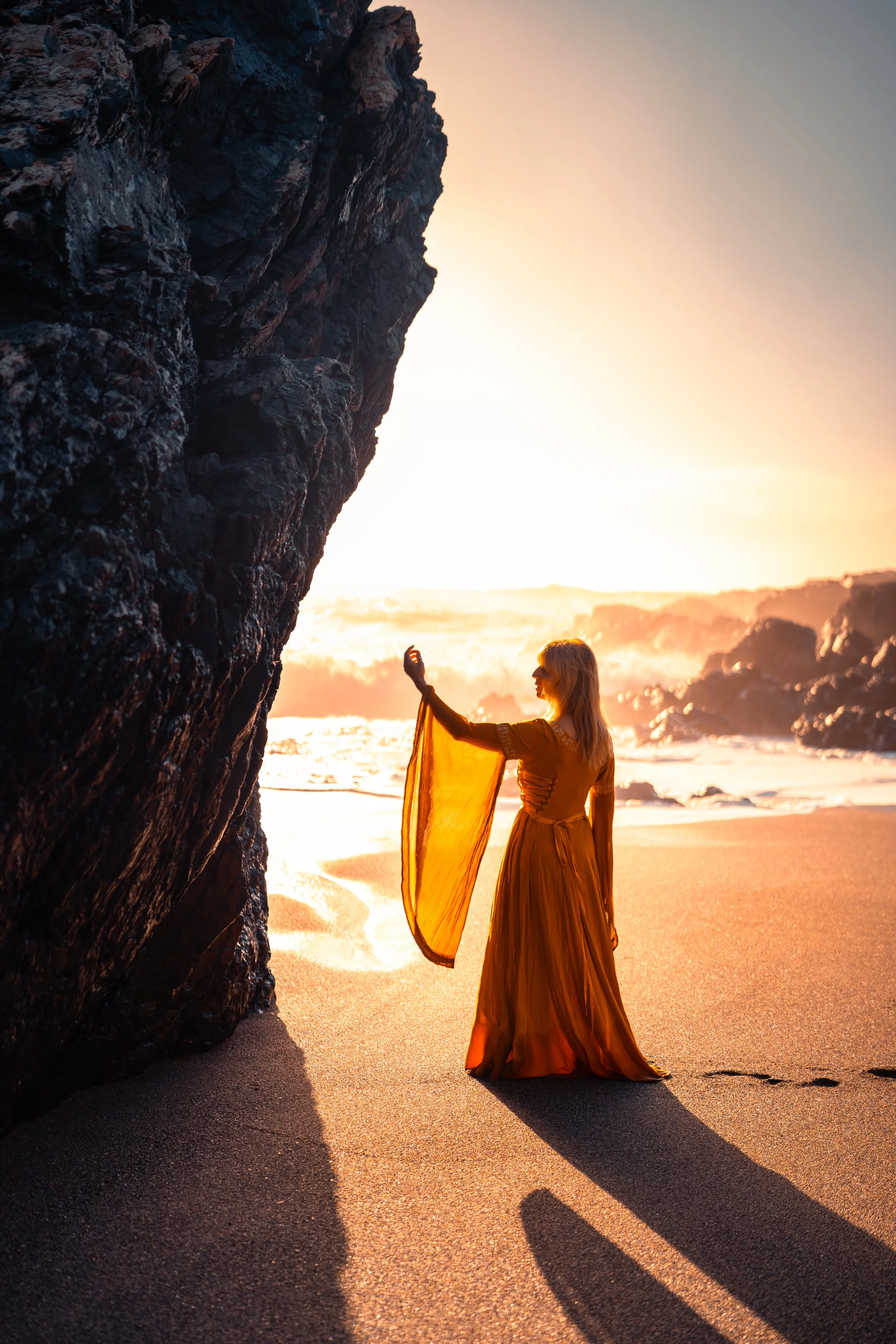 A woman in a flowing dress standing on a beach at sunset near a large rock formation, with the sun casting a golden glow over the scene.