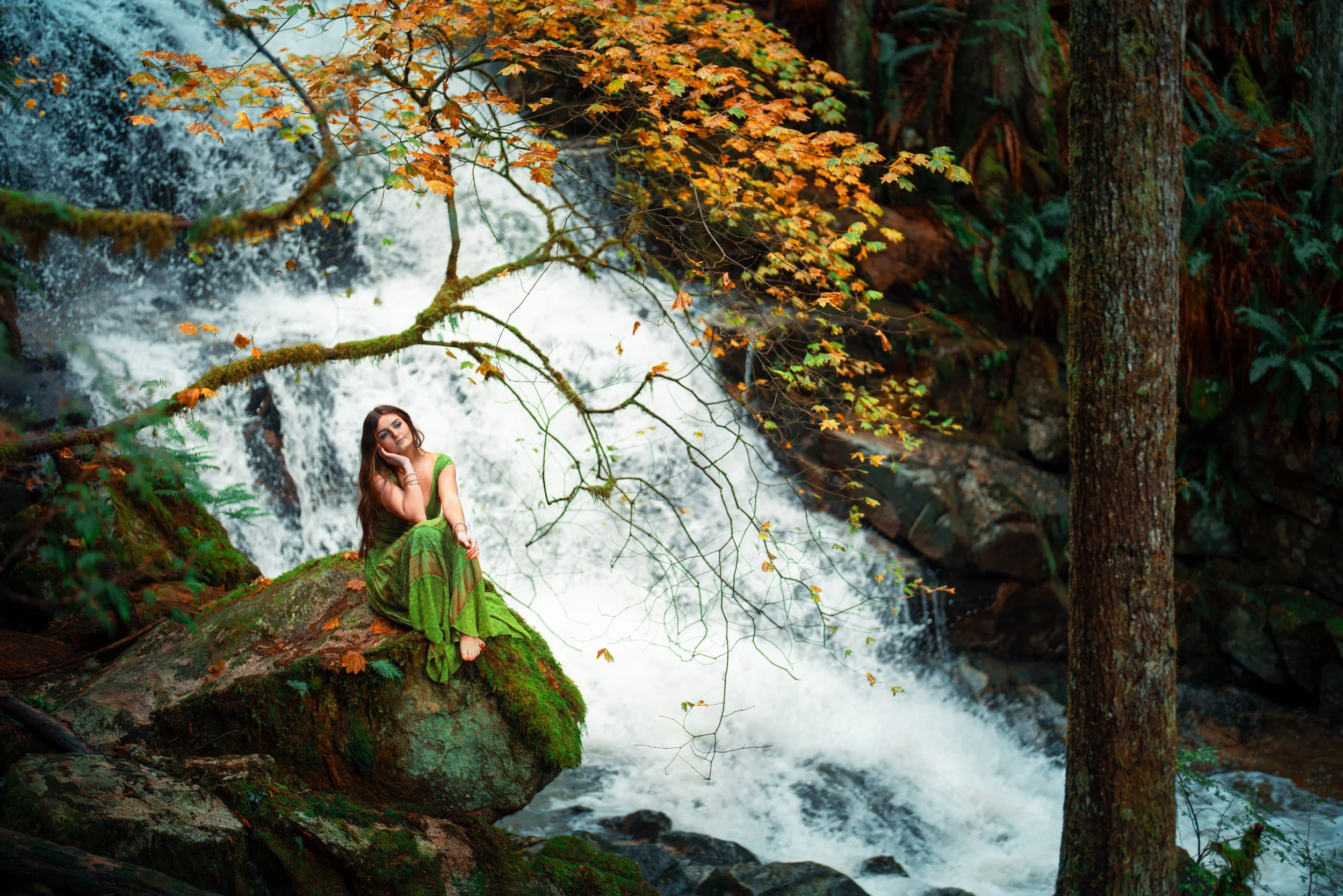 A woman wearing a green dress sitting on a moss-covered rock in front of a waterfall in a forest with trees and orange leaves.