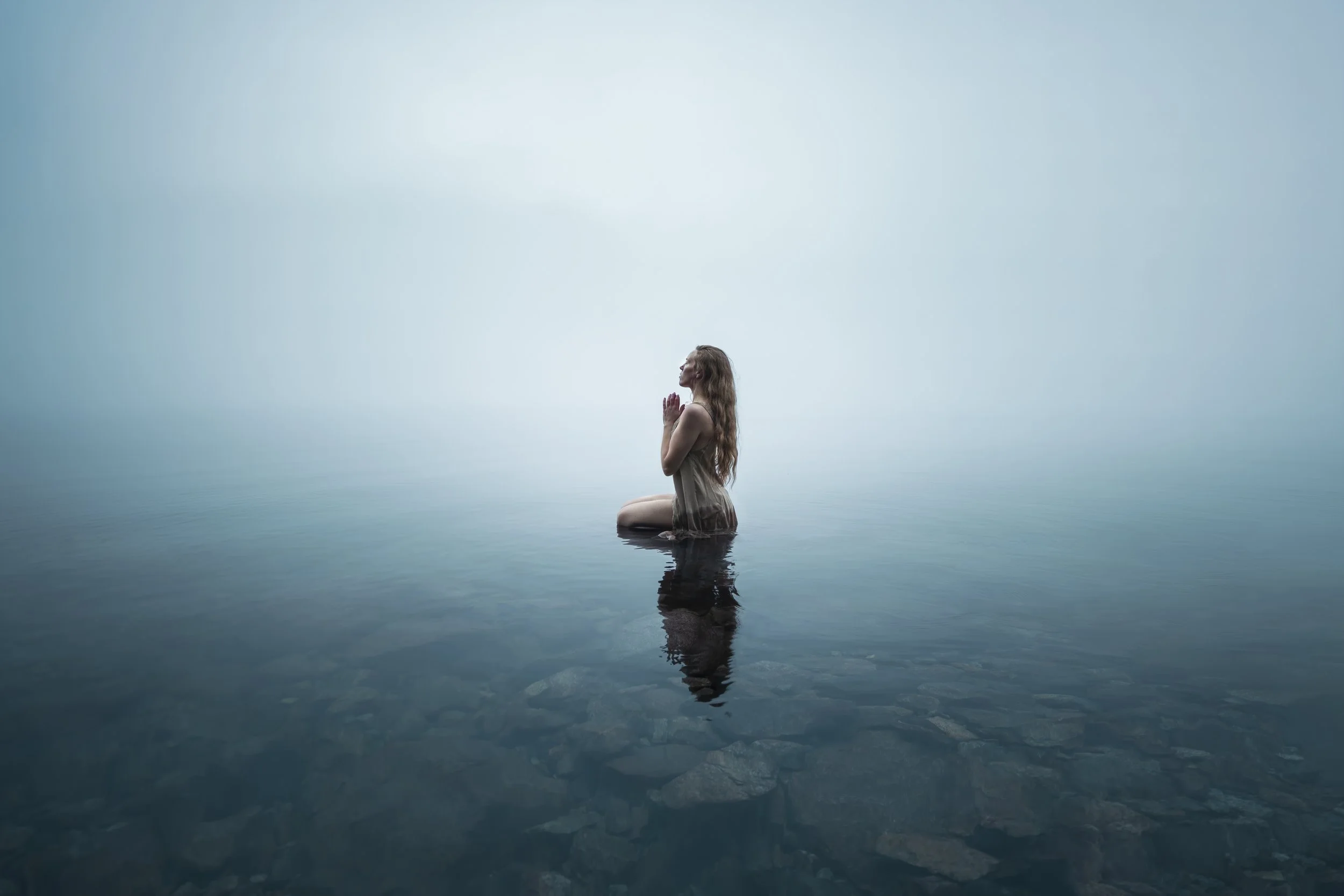 A woman kneeling in calm water with her hands clasped in prayer, facing upward, with a foggy or misty background.