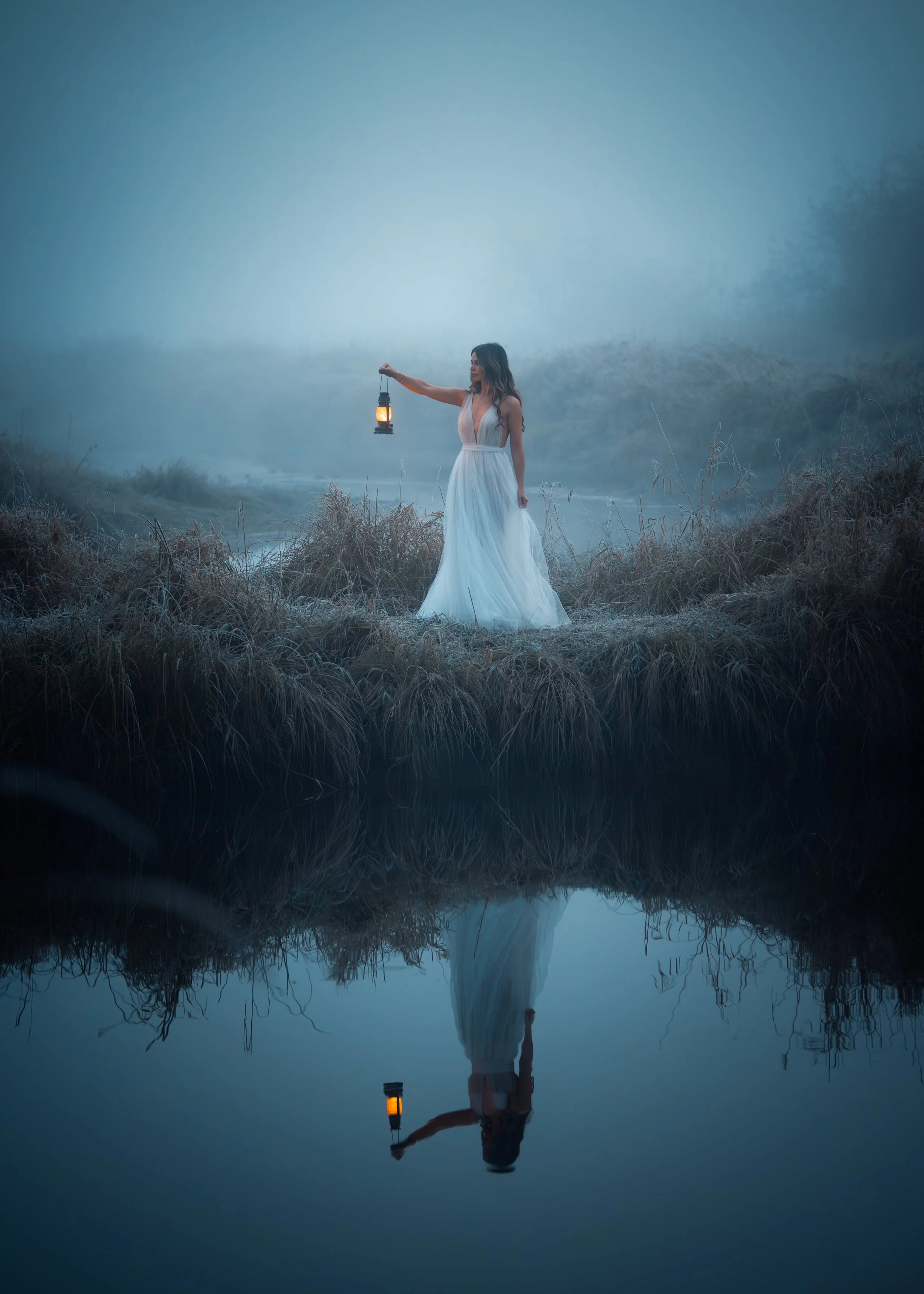 A woman in a white dress holding a lantern stands on a grassy bank near water with foggy trees in the background. Her reflection is visible in the water.