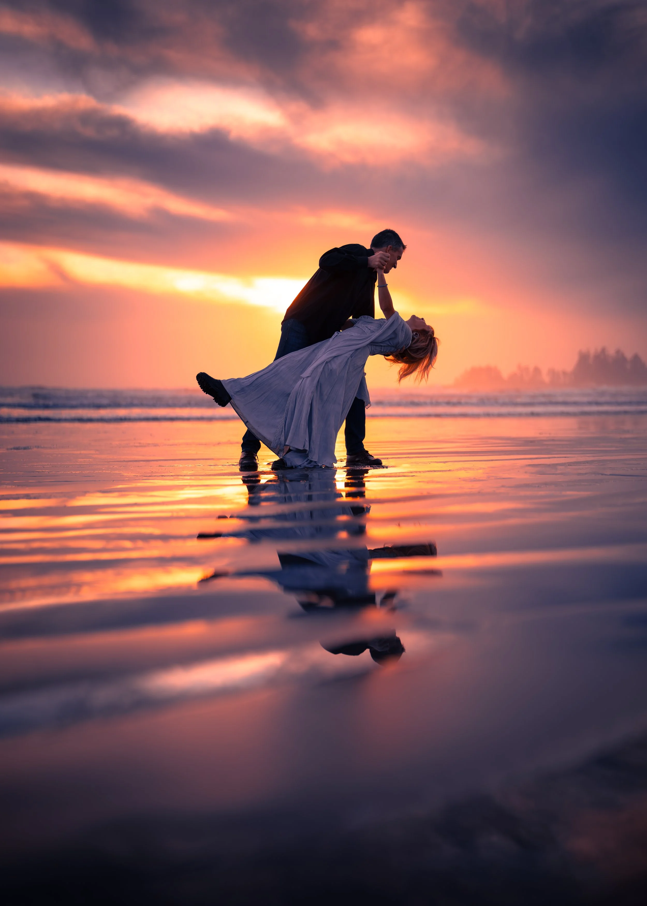 A couple dancing on the beach during sunset with a colorful sky and reflections on the wet sand.