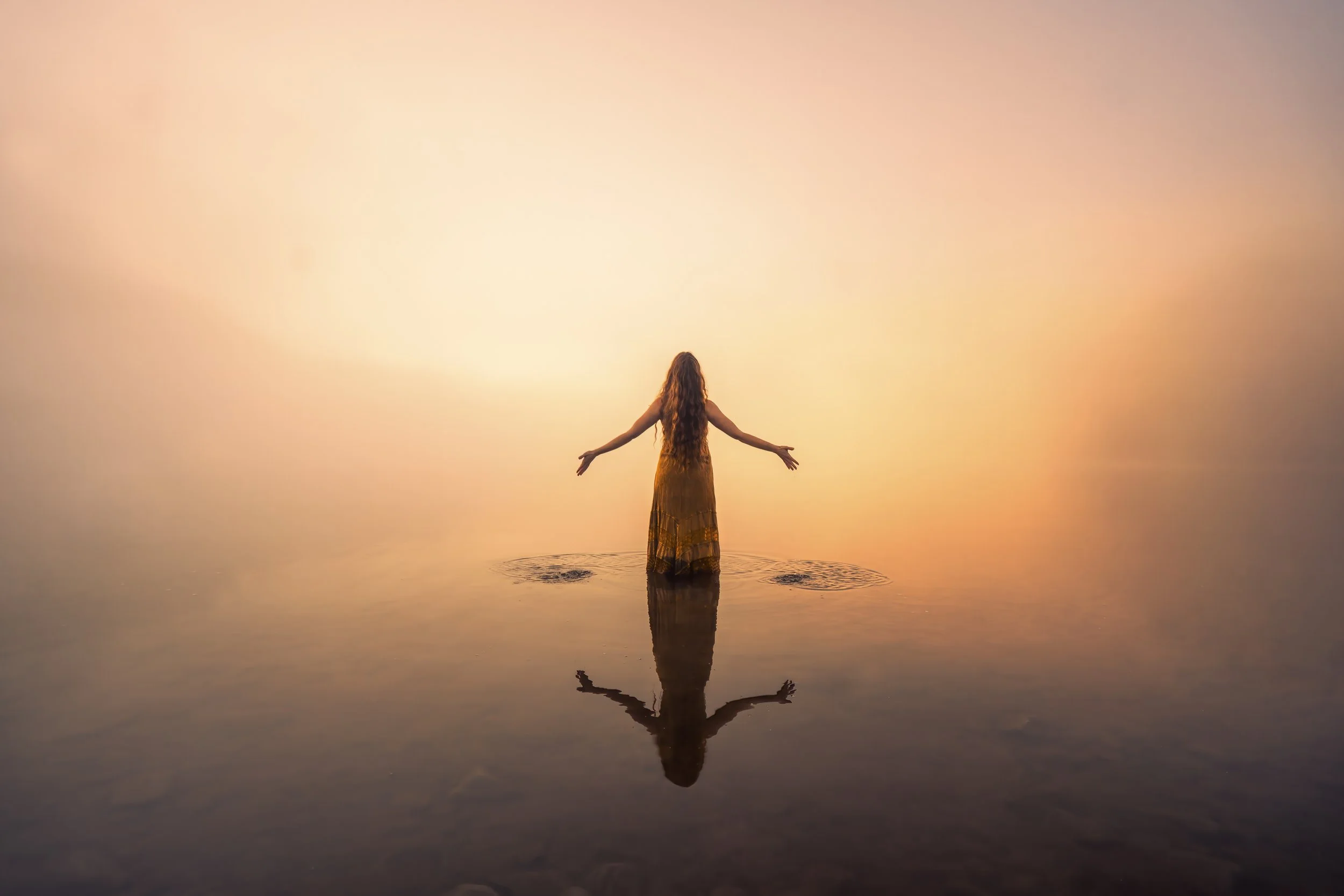 A woman with long hair standing in calm water during sunset or sunrise with arms outstretched.