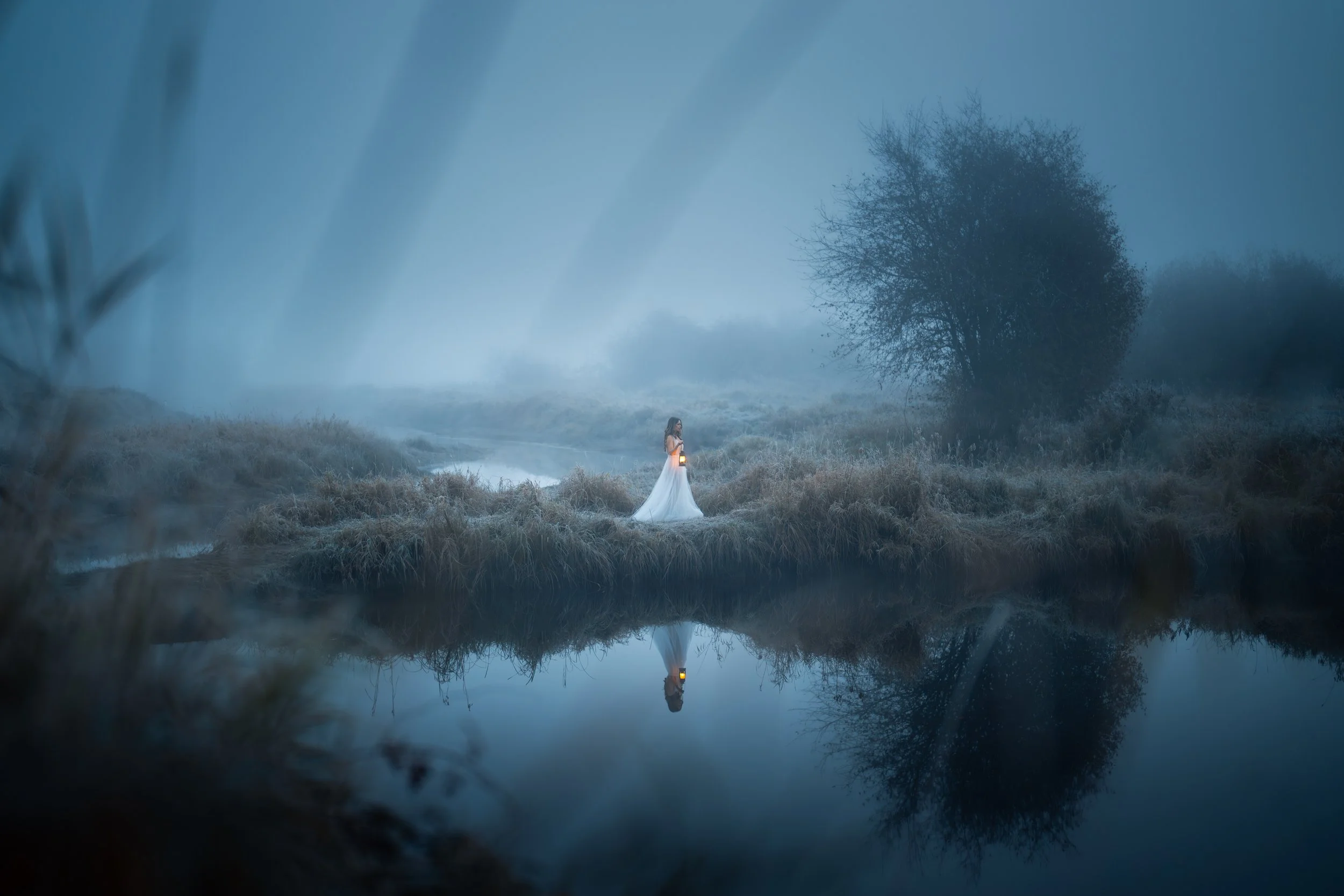 A woman in a white dress holds a lantern while standing on a grassy riverbank in a foggy, wooded area, with her reflection visible in the still water.