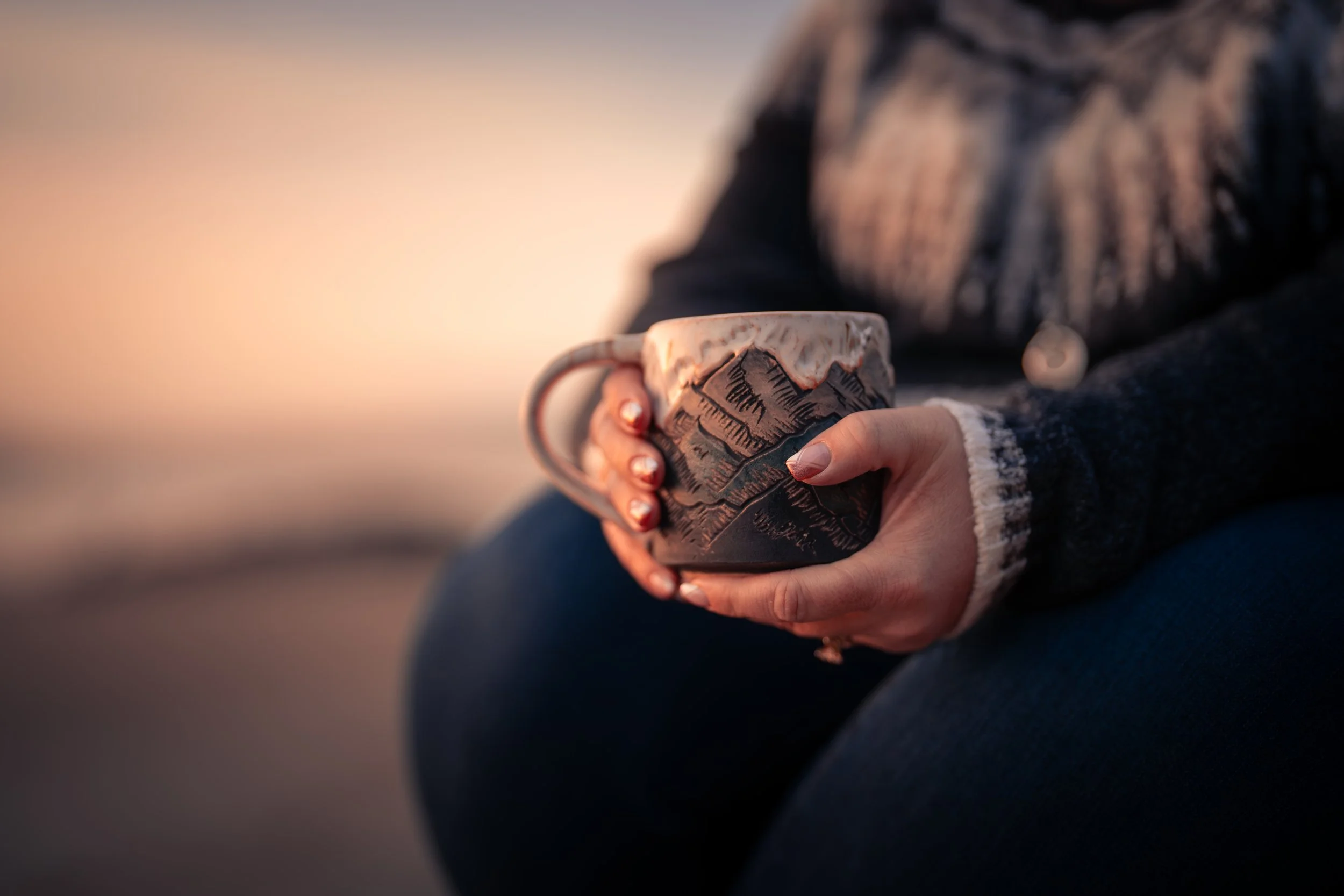 Person holding a mug decorated with mountain landscape design, sitting outdoors at sunset.