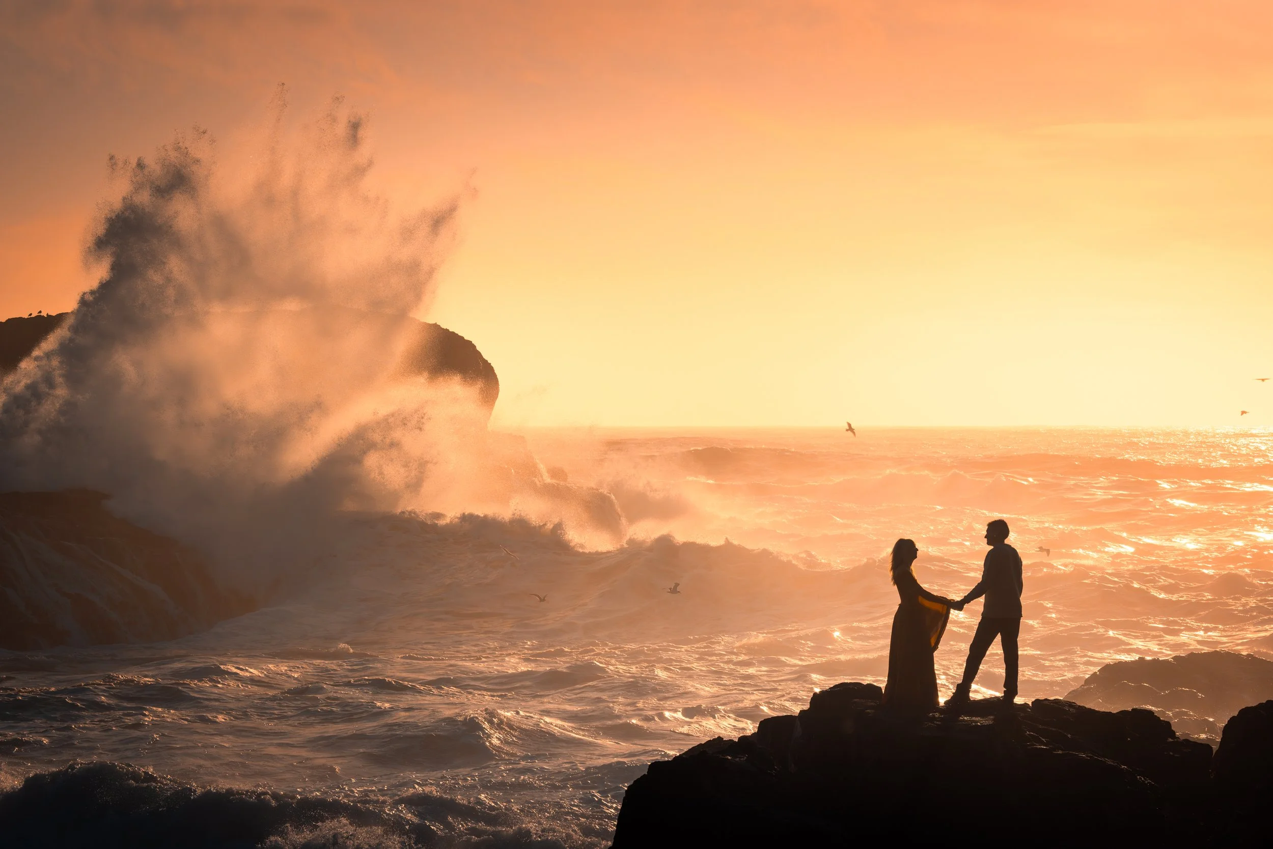 A couple standing on rocks holding hands during sunset by the ocean, with waves crashing and seagulls flying.