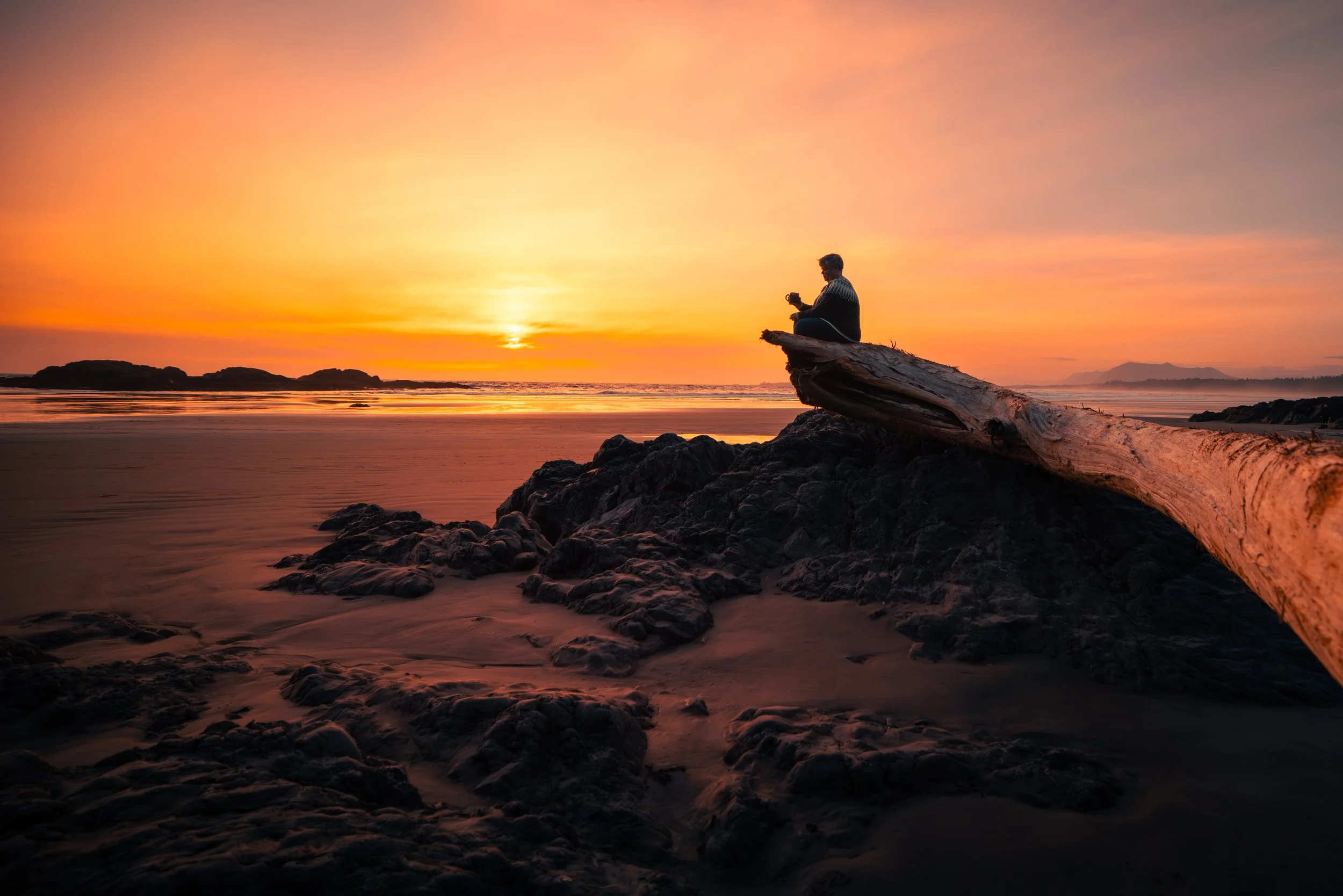 Person sitting on a large piece of driftwood on a rocky beach at sunset, with the ocean and distant islands in the background.