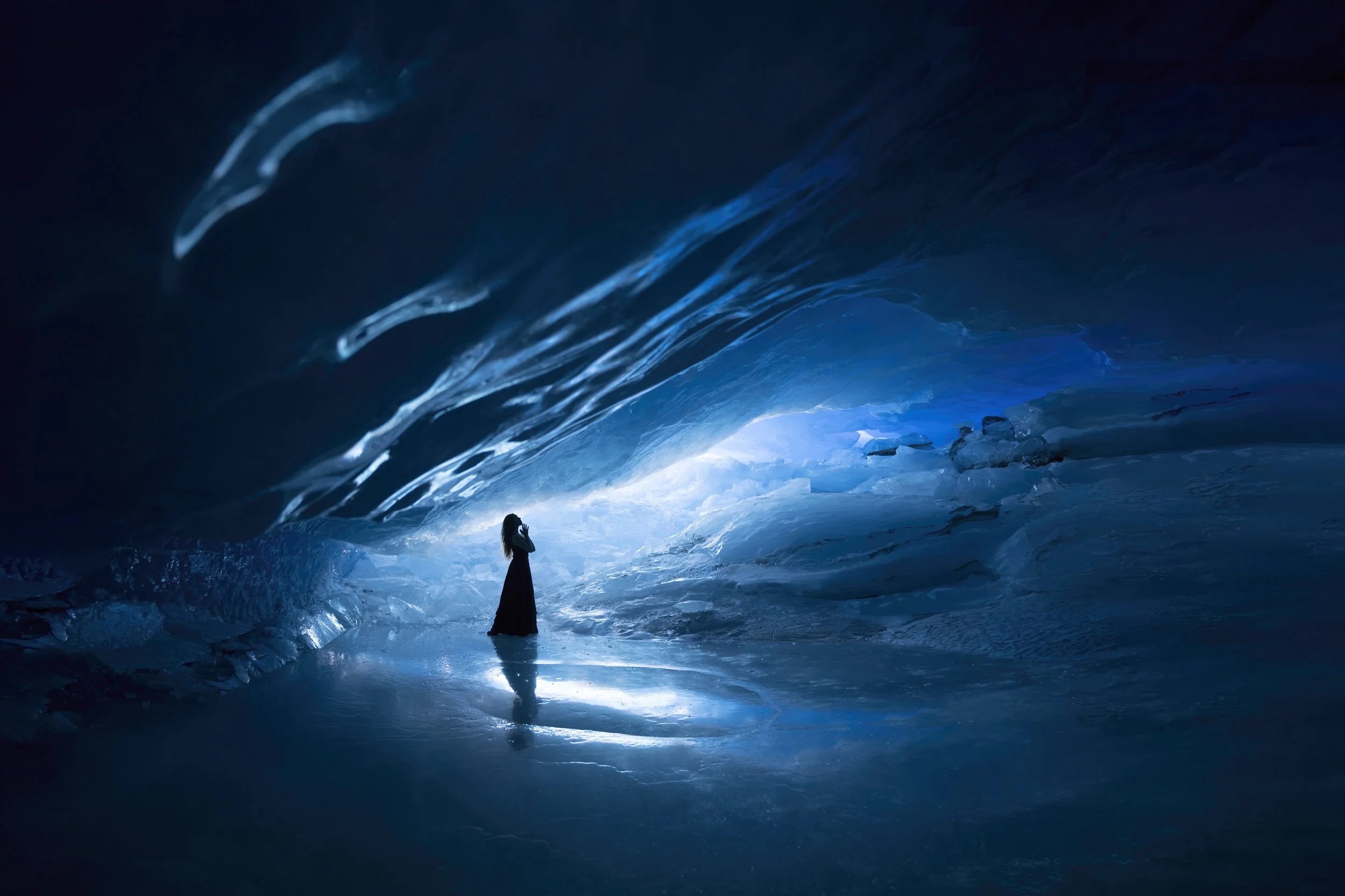A woman standing inside an icy glacier cave, illuminated with blue light, with icy walls and a reflective ice floor.