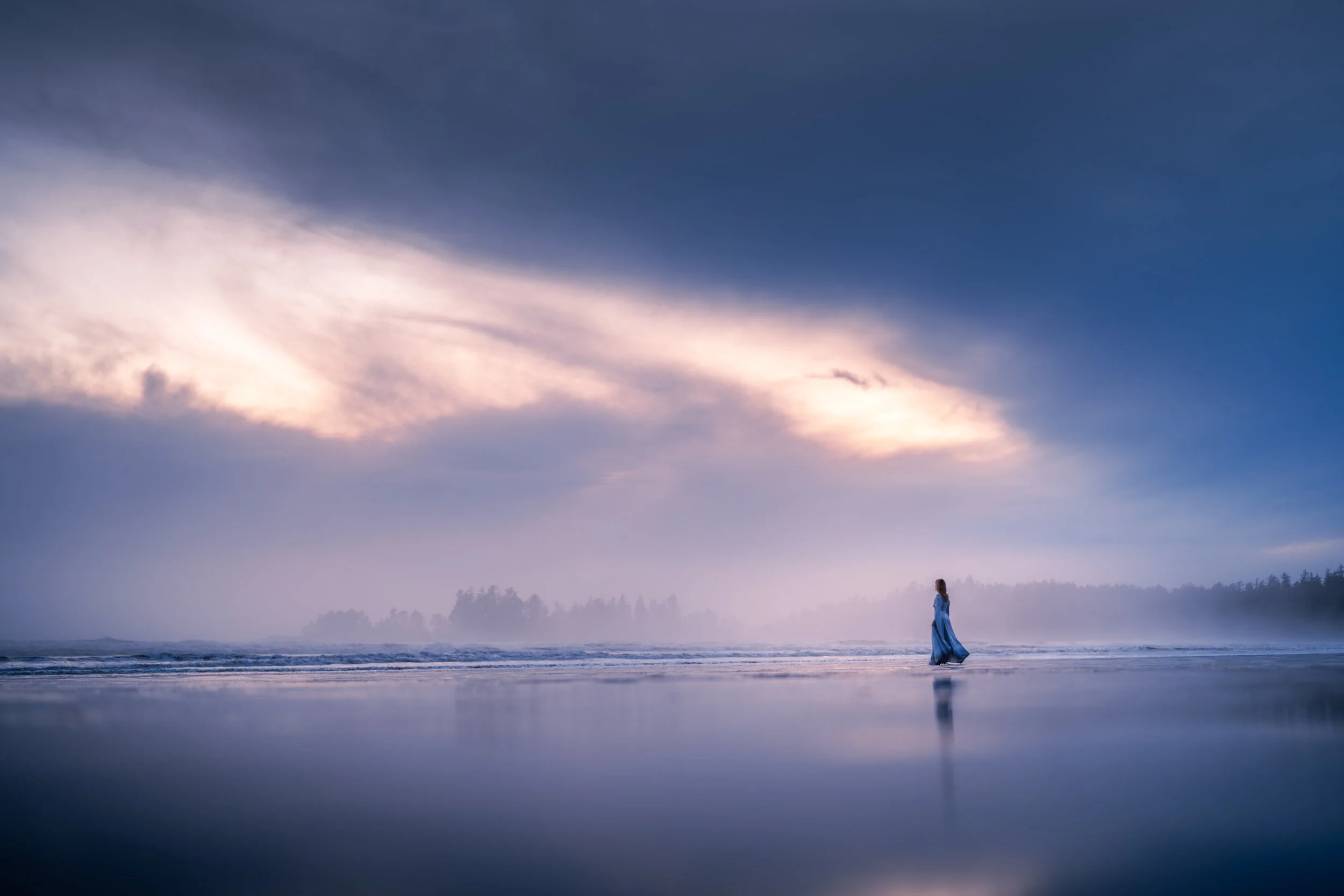A woman in a blue dress walking along the shoreline at sunset, with a colorful sky and distant trees in the background.