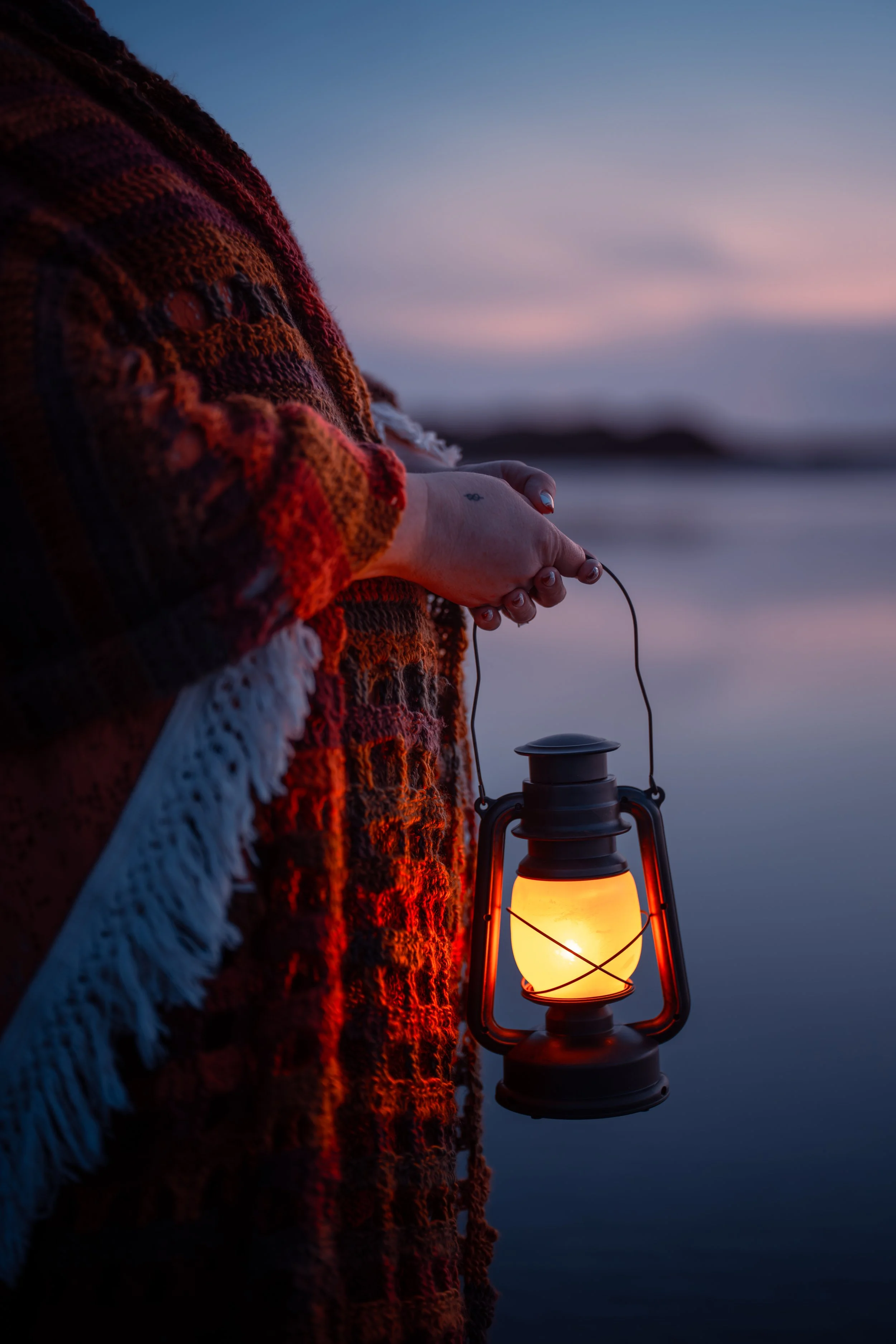 Person wearing a warm, colorful sweater holding a lantern near a body of water during twilight.