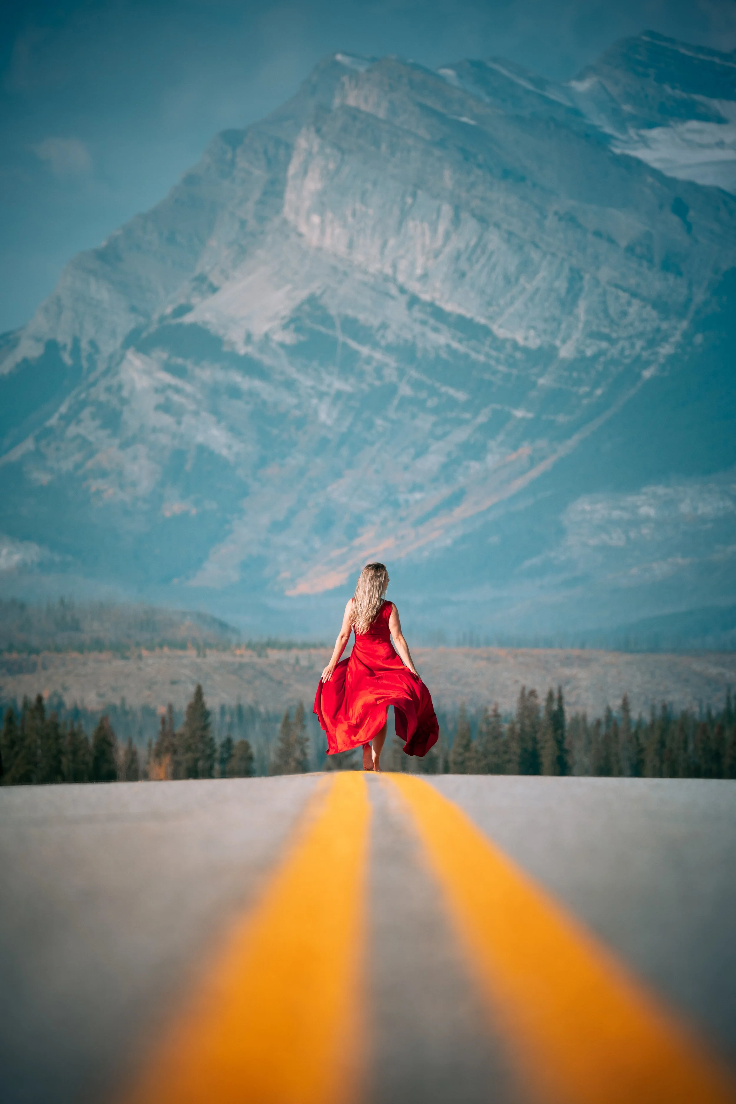 A woman in a red dress walking alone on a deserted mountain road with a large mountain in the background.