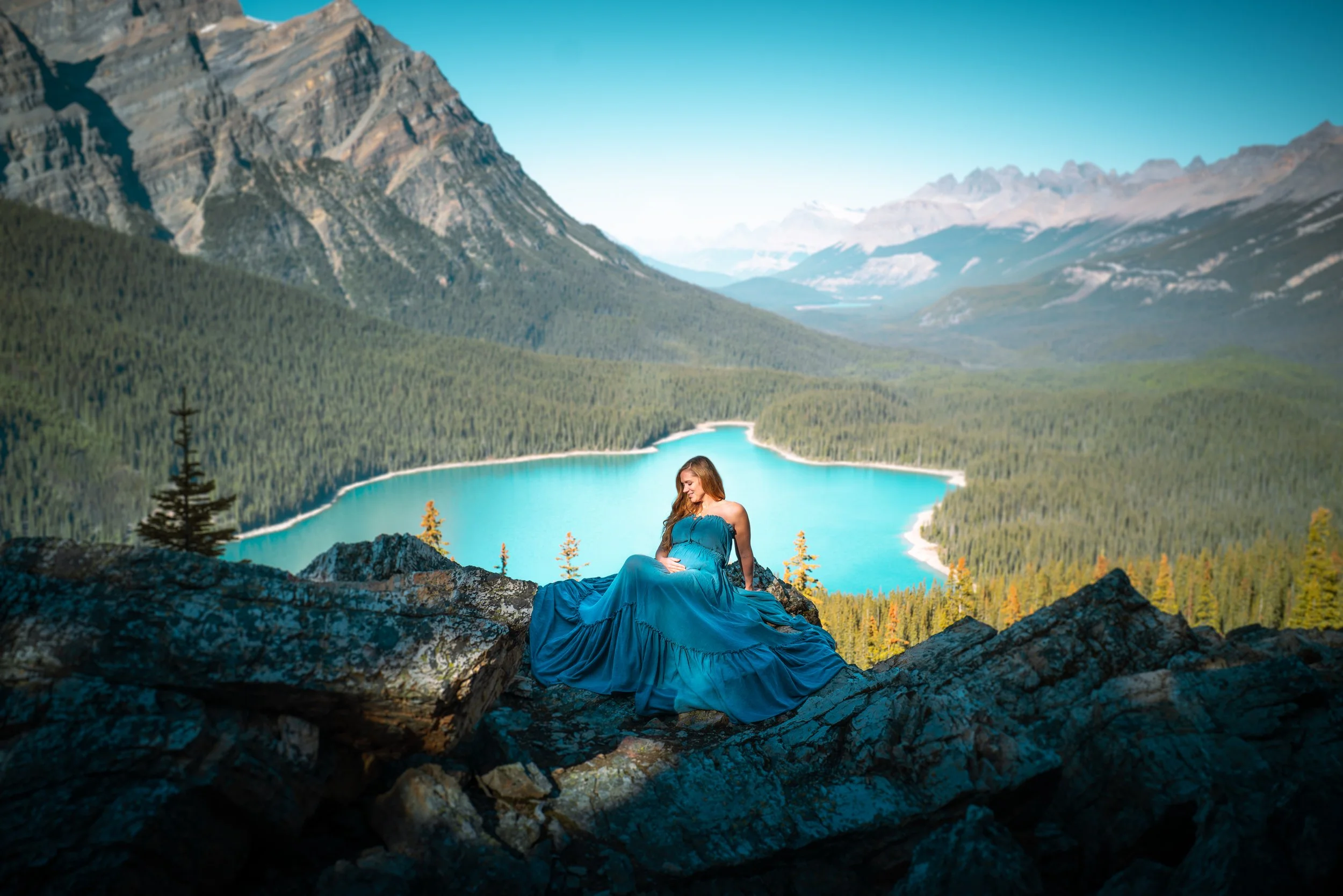 A woman in a blue dress sitting on rocks overlooking a mountain lake surrounded by forest.