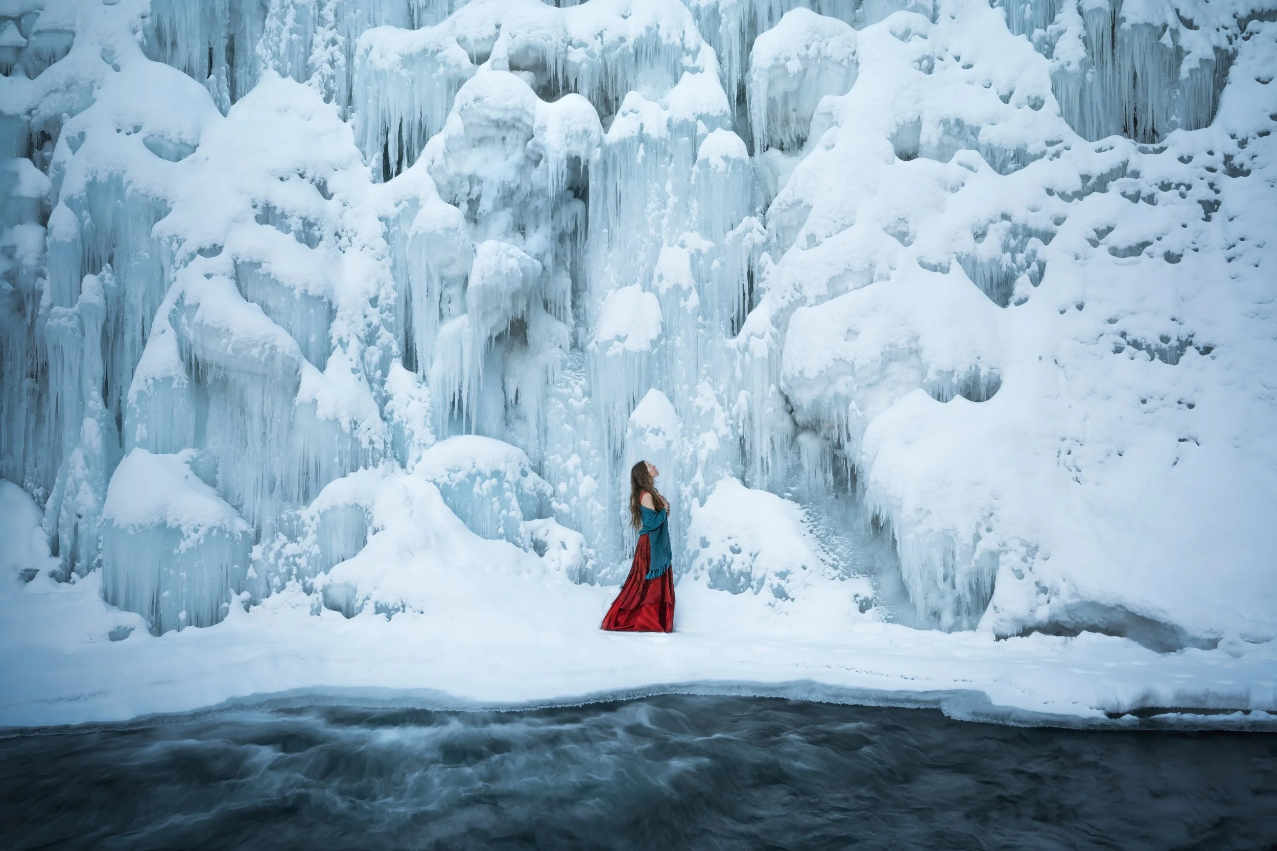 A woman in a blue and red dress stands on snow-covered ground near a large ice wall with icicles and snow, next to a body of dark water.