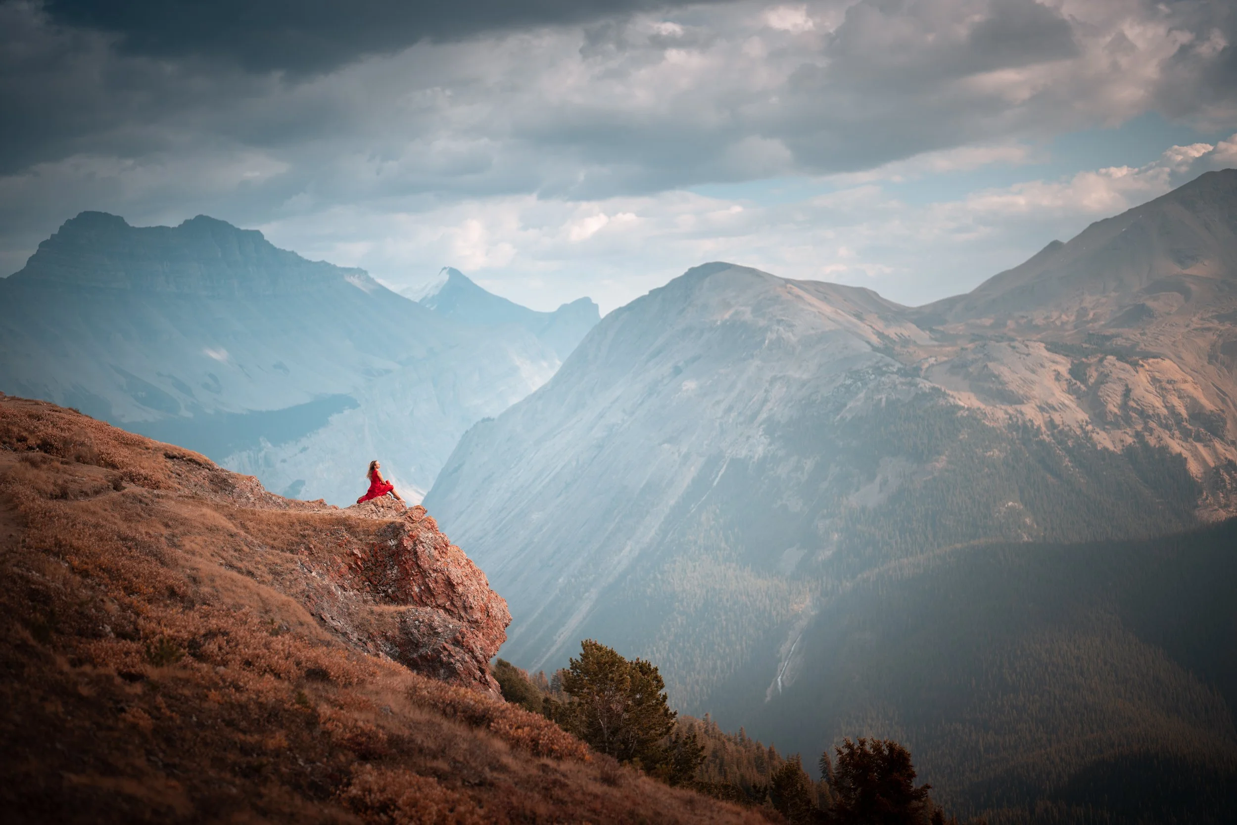 A person in red sitting on a rocky outcrop overlooking a vast mountain landscape with peaks, valleys, and a cloudy sky.
