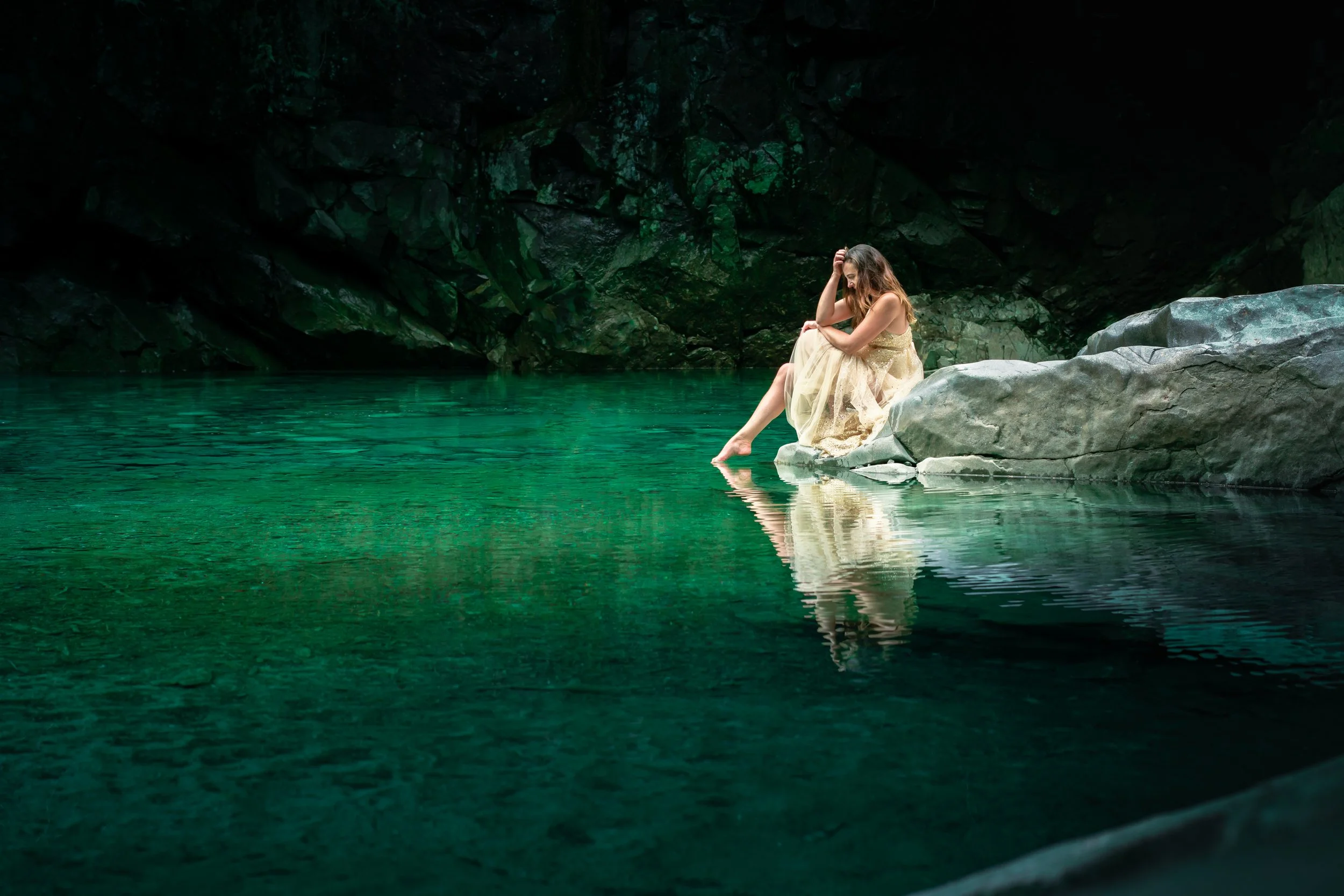 A woman in a light-colored dress sitting on a rock by a calm, greenish body of water in a dark cave.