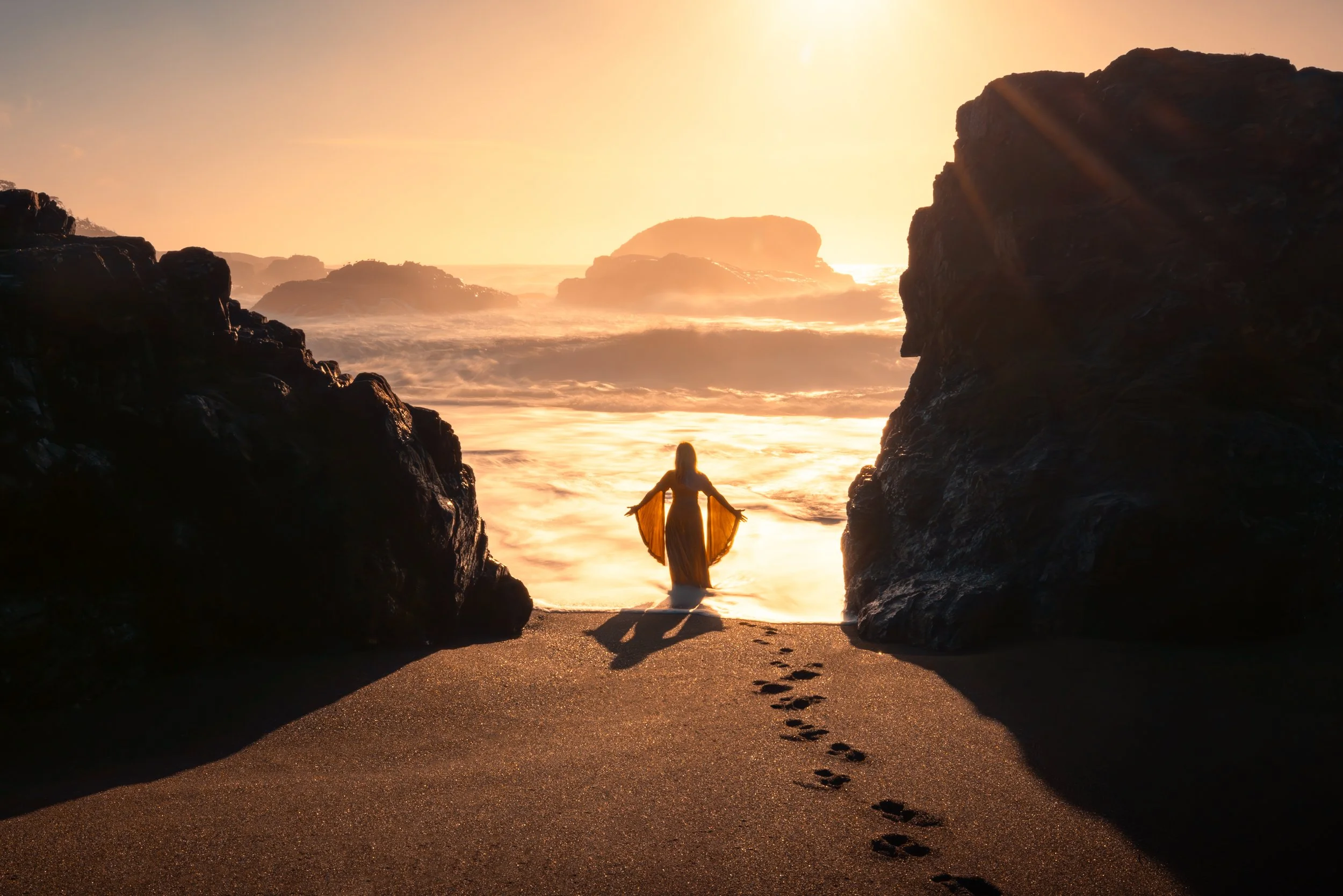 A woman in a flowing dress standing on a beach at sunset, framed by large rocks, with footprints in the sand leading to her.