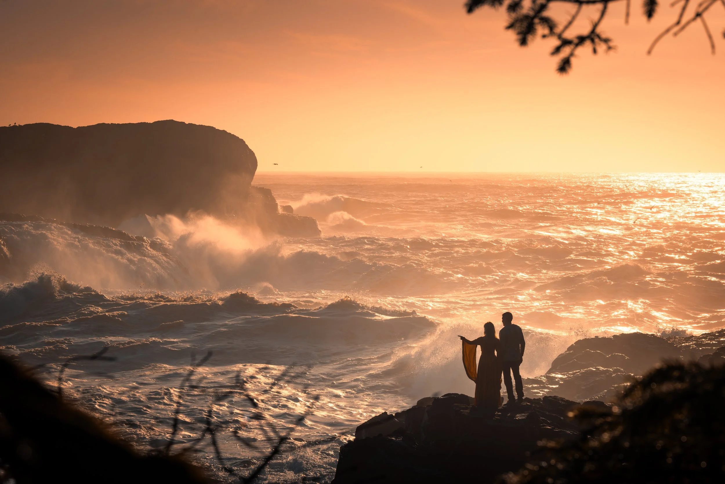 Silhouettes of a man and woman standing on rocks by the ocean during sunset, with waves crashing and a cliff in the background.