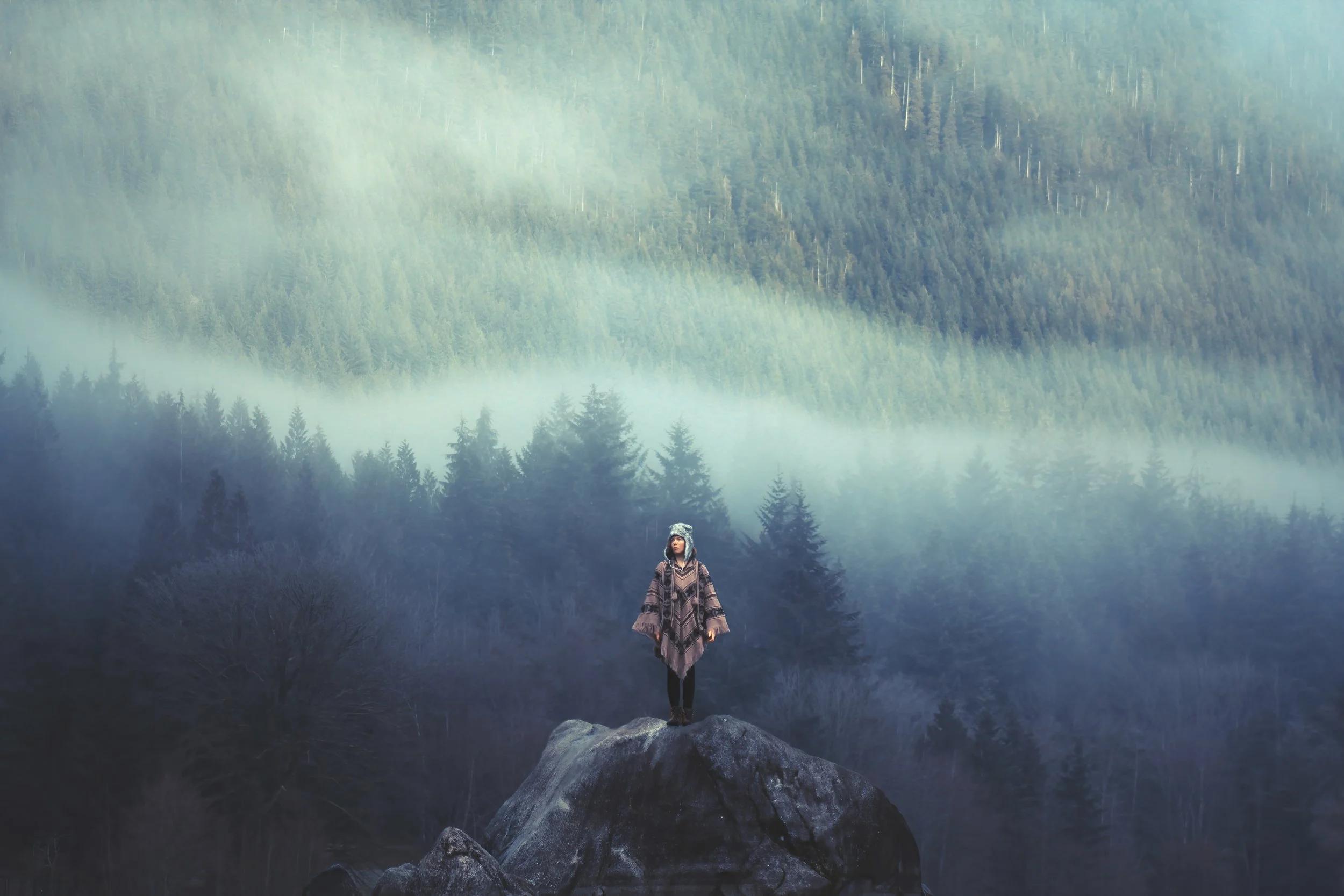A woman standing on a large rock in a forested mountainous area with fog and trees in the background.