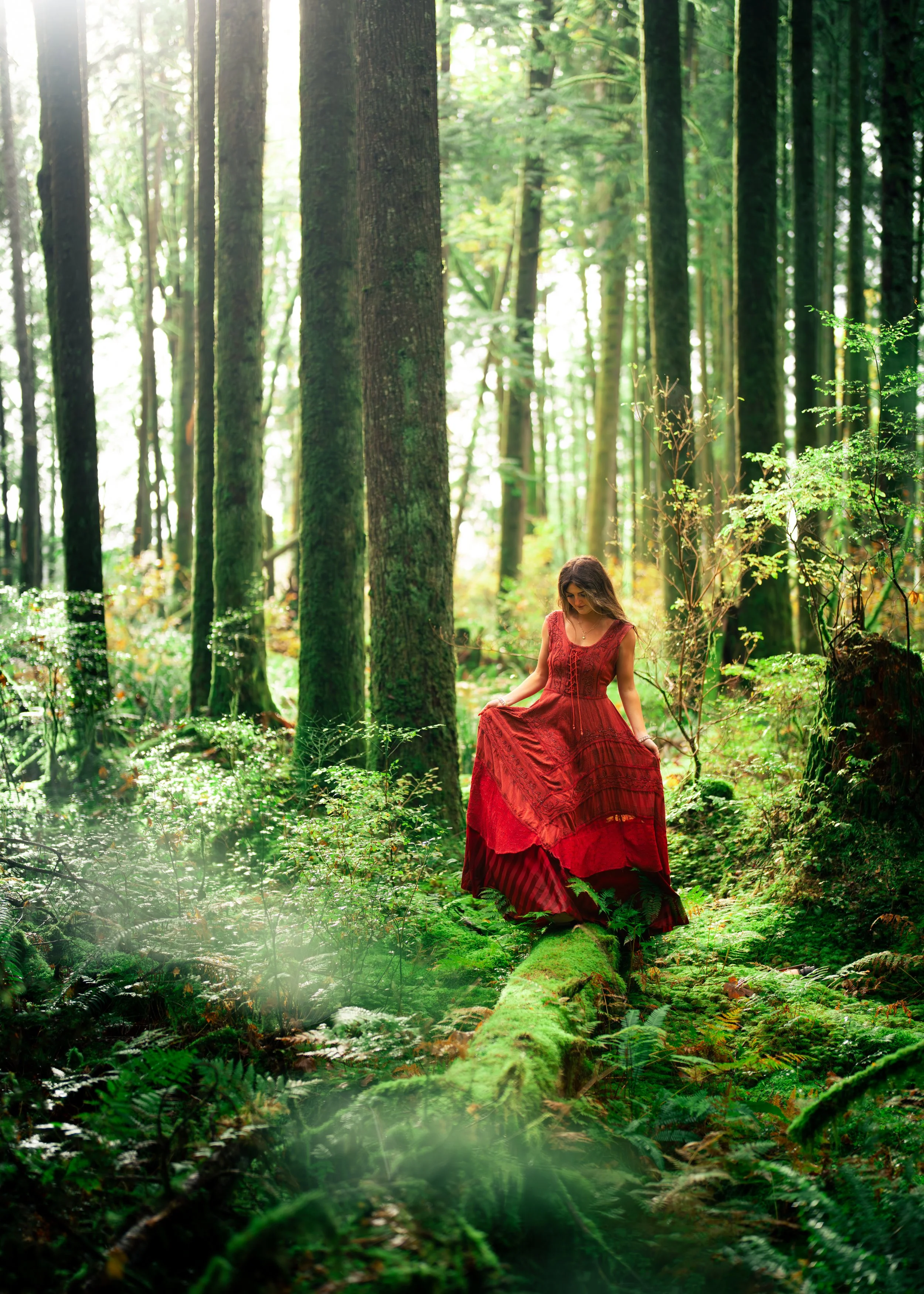 A woman in a long red dress standing on a moss-covered fallen tree in a lush green forest with tall trees and dense foliage.