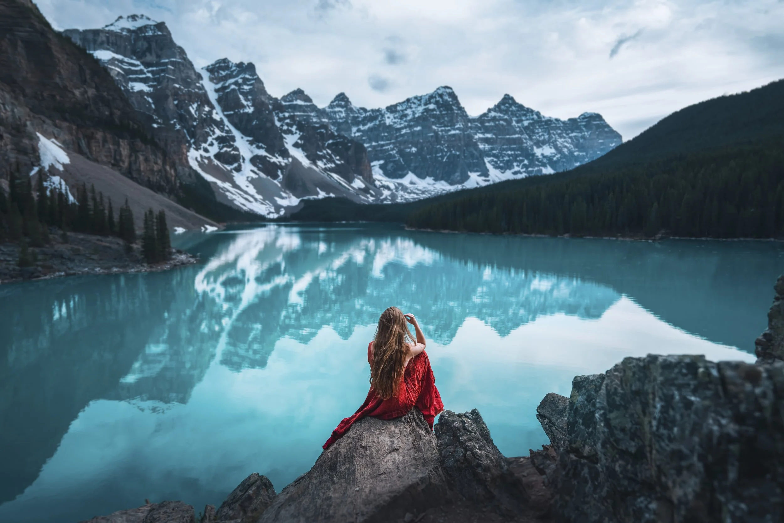 A woman in a red dress sitting on rocks by a turquoise lake with snow-capped mountains and evergreen trees in the background.