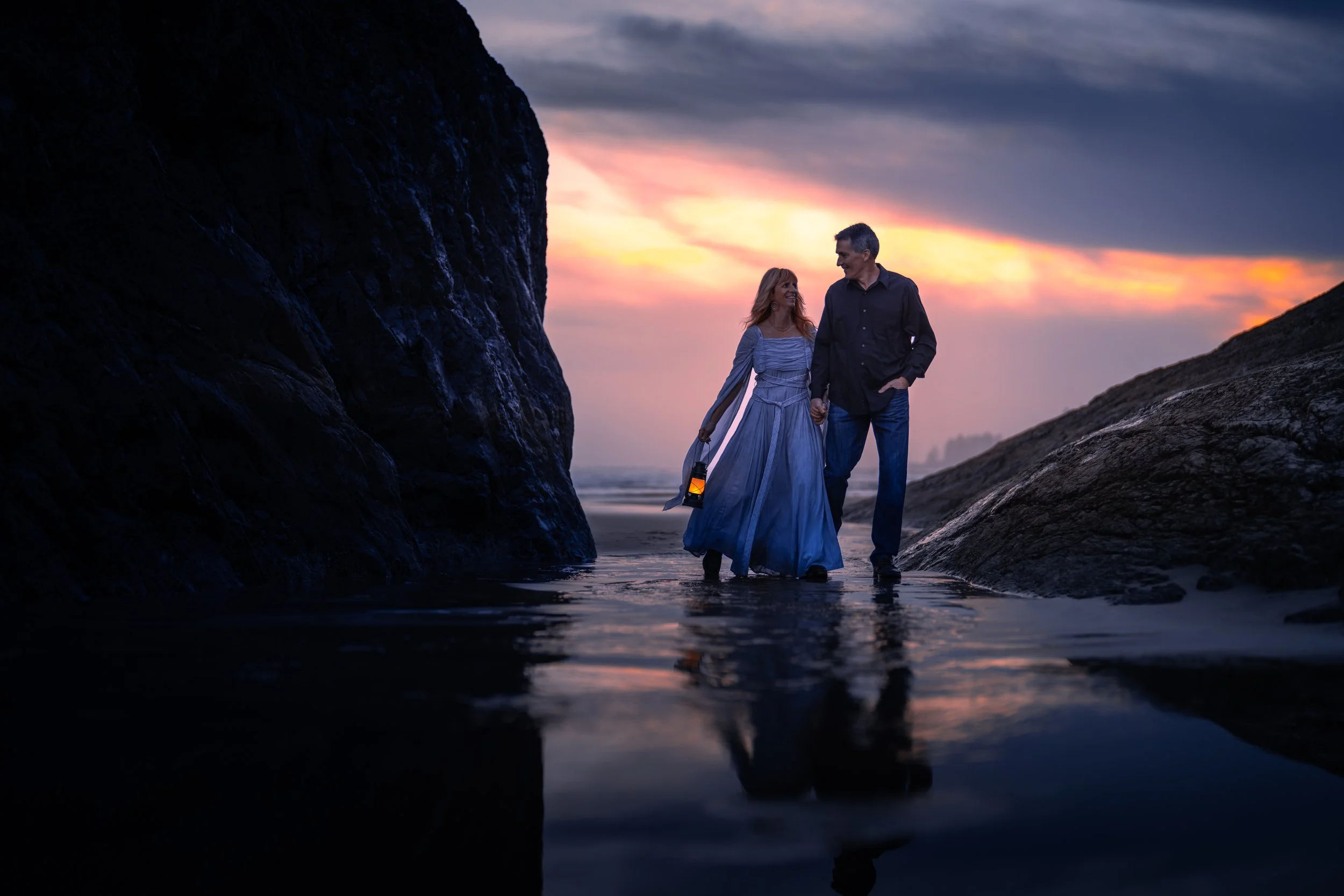 A couple walking hand in hand on a beach at sunset, with rocky formations on either side and the sky painted in shades of orange and purple.