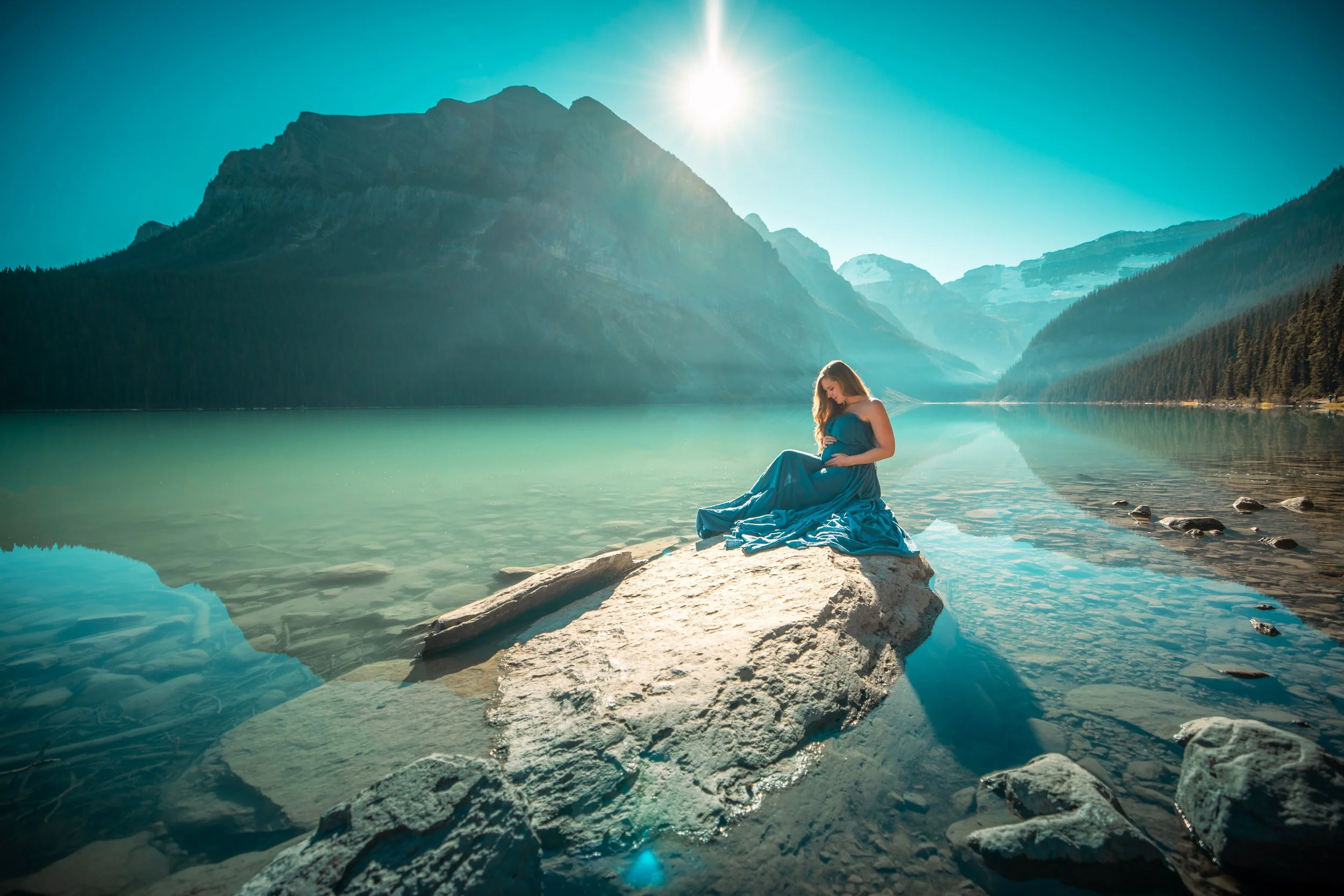 A woman in a blue dress sitting on a large rock by a calm lake with mountains in the background and the sun shining brightly overhead.