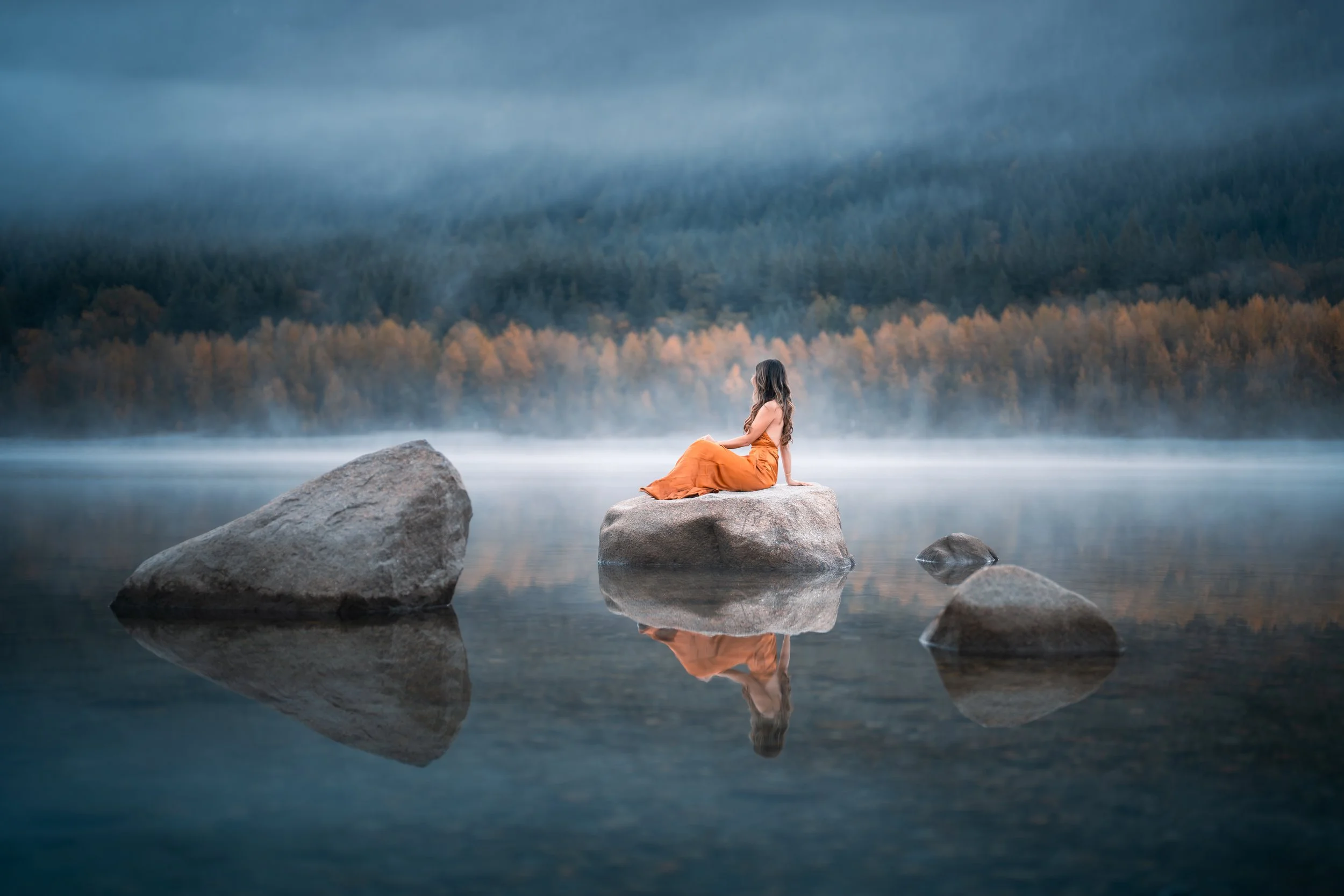 A woman sitting on a rock in the middle of a calm lake with mist, surrounded by mountains and a forest, during foggy weather.