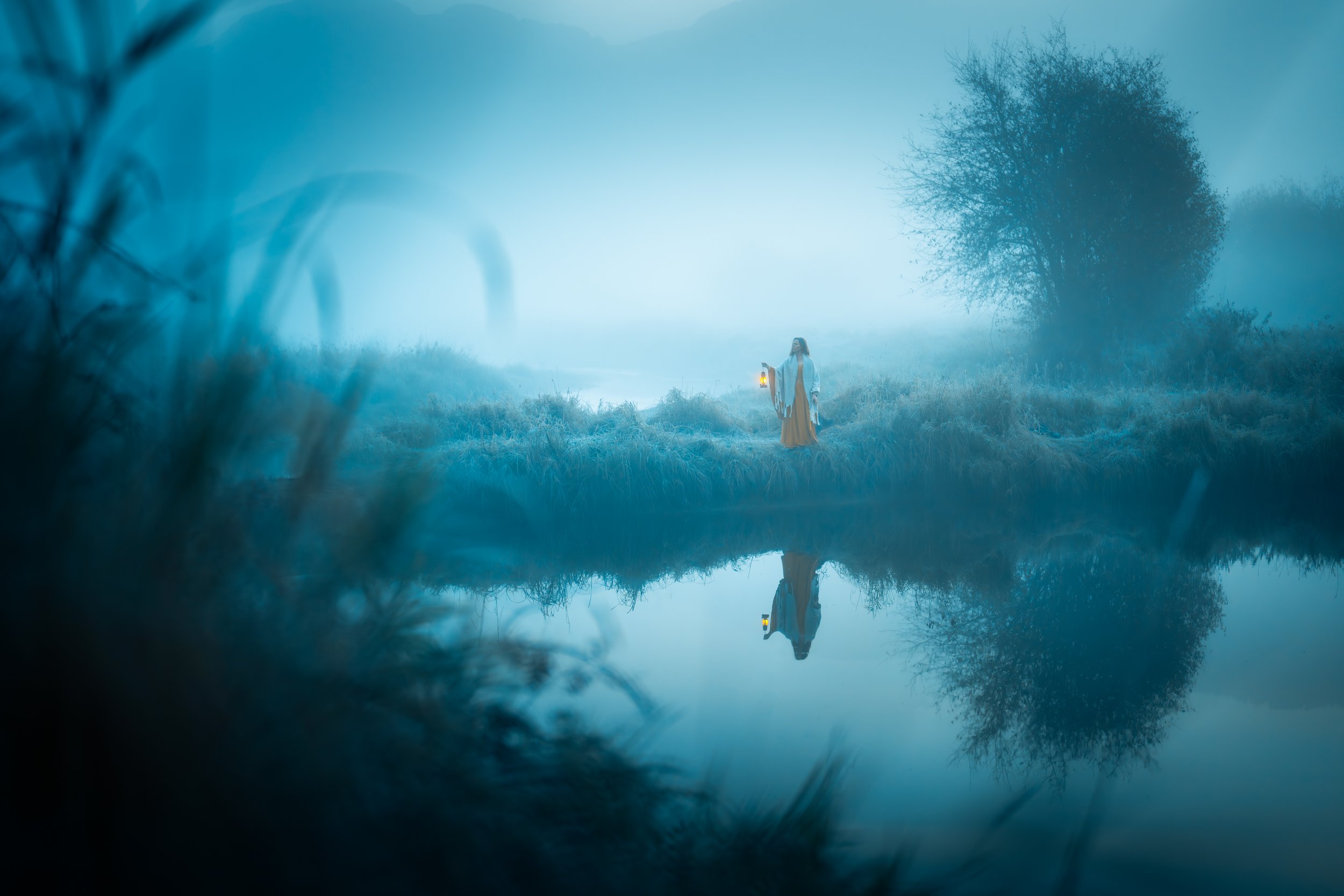 A person dressed in historical medieval clothing holding a lantern, standing by a foggy riverbank with trees in the background, their reflection visible in the water.