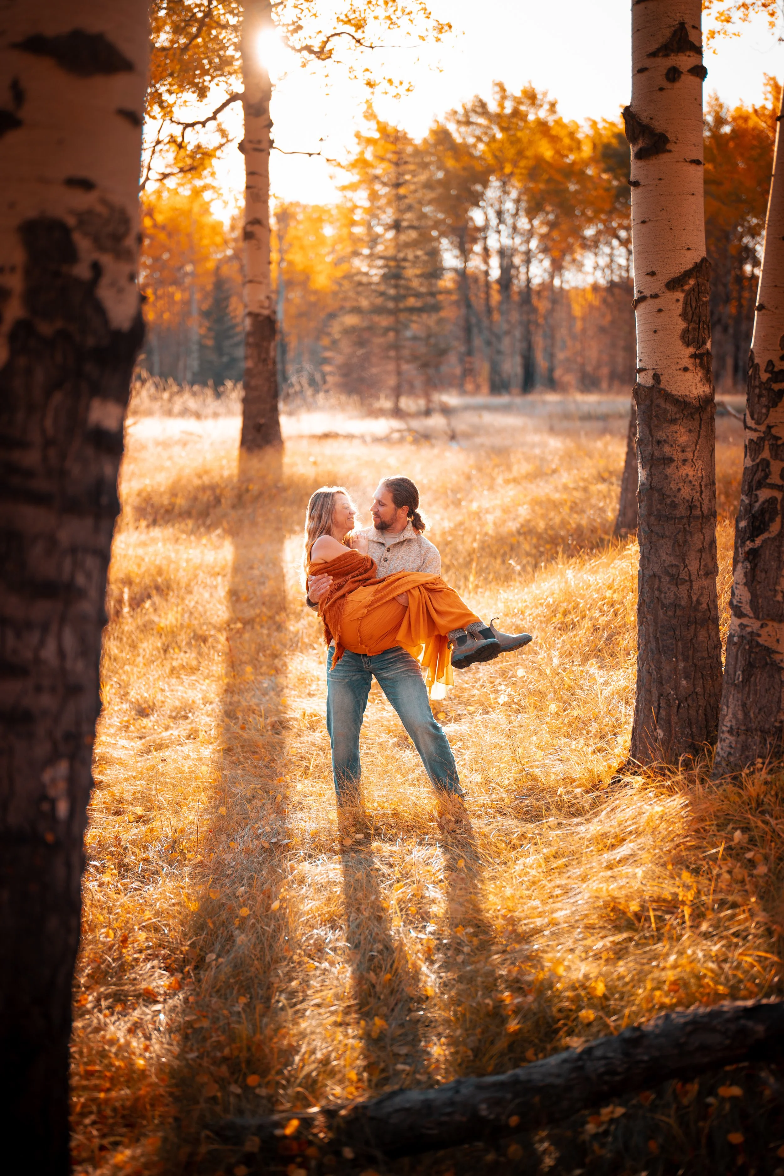 A man holding a woman in his arms in an autumn forest with tall trees and golden leaves, sun shining through the trees.