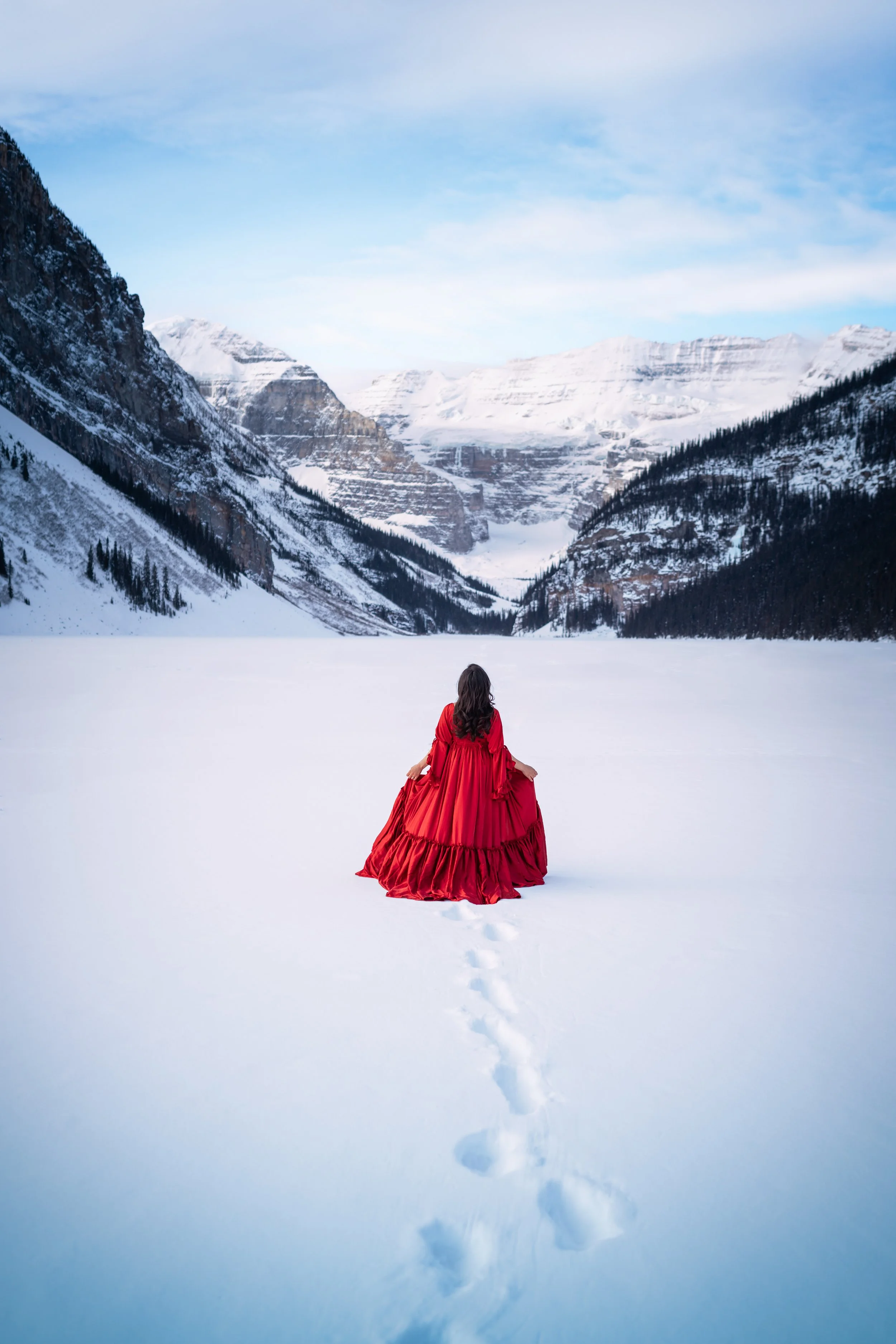 A woman in a red dress sitting on snow in a snowy mountain valley with tall snow-covered peaks and a blue sky in the background.