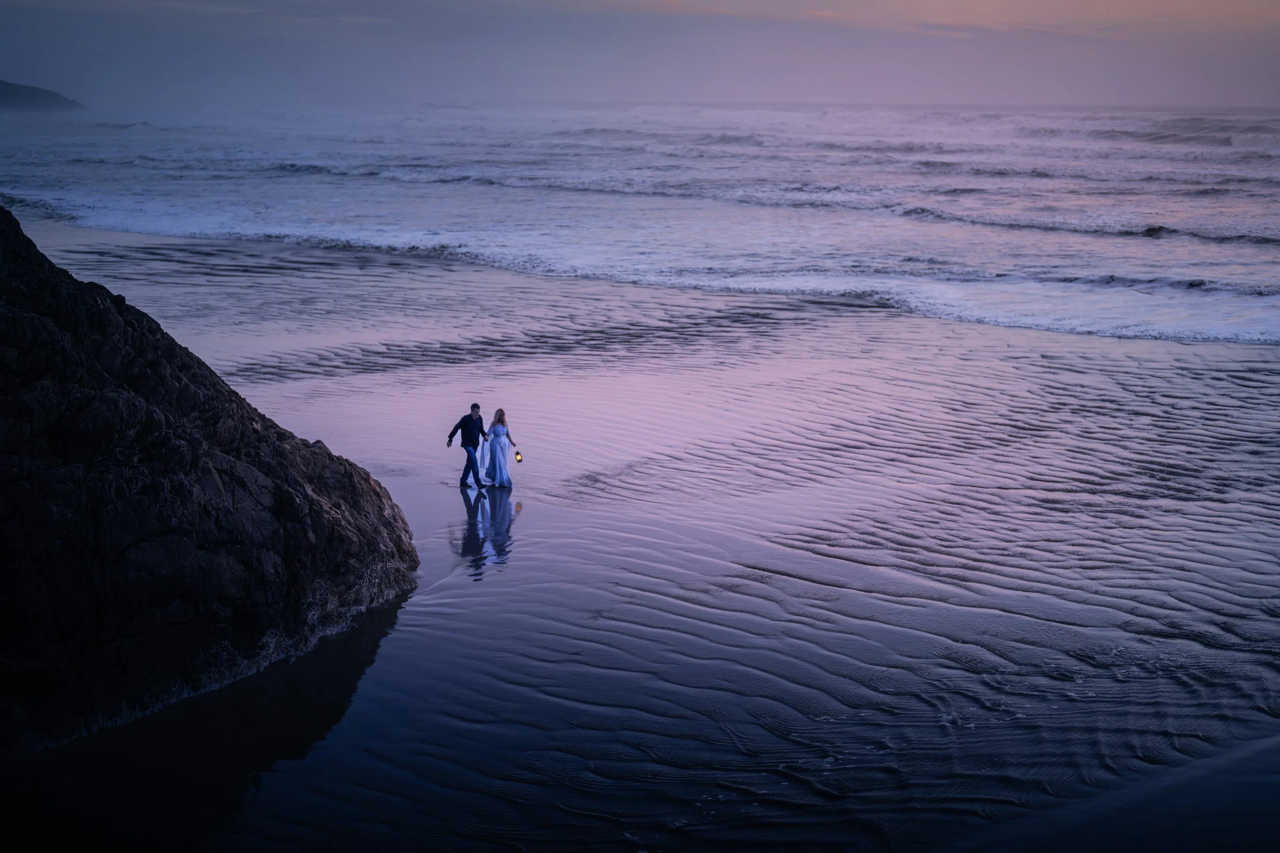 A couple walking along the beach at sunset with the woman in a long blue dress holding a lantern, waves gently crashing on the shore, reflecting purple and pink hues in the water.