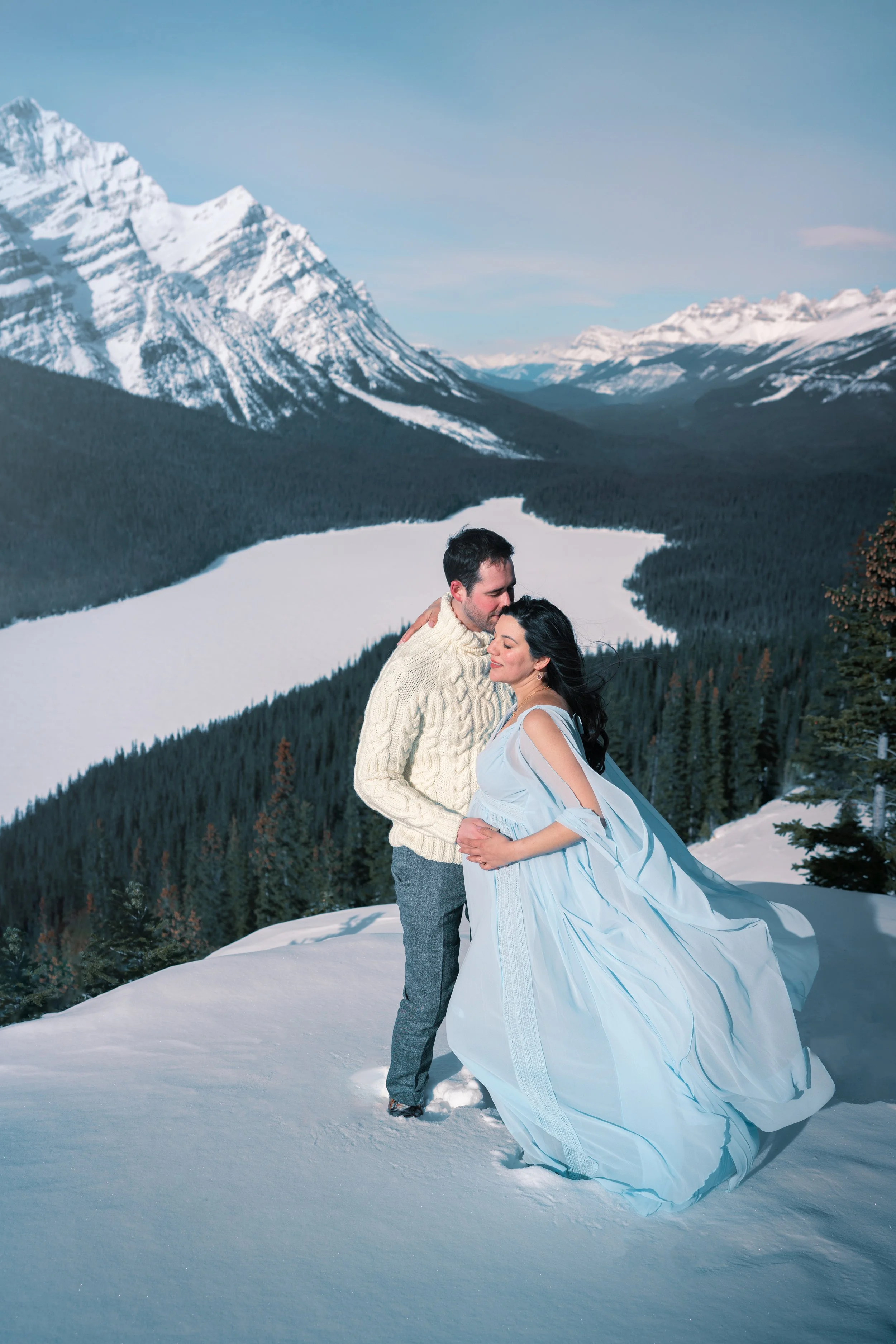 A couple in wedding attire standing on snow-covered ground in a mountainous landscape with snow-capped peaks, a river or lake, and pine trees in the background.