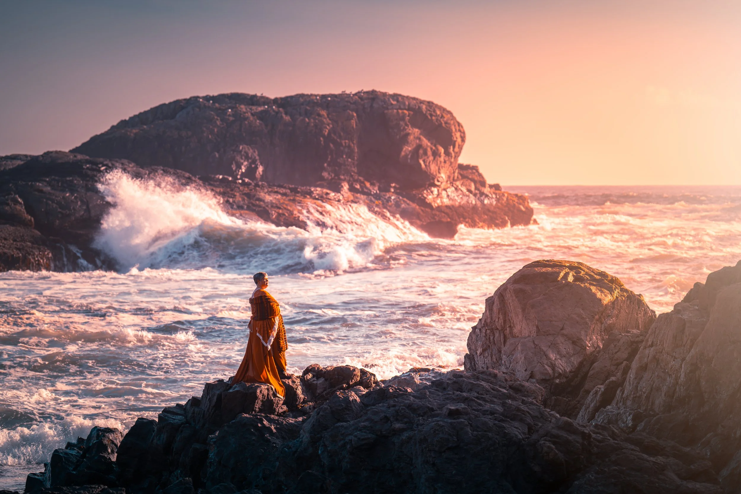 A woman in a long orange dress stands on rocks by the ocean at sunset, facing the waves crashing on the shore with a large rocky hill in the background.