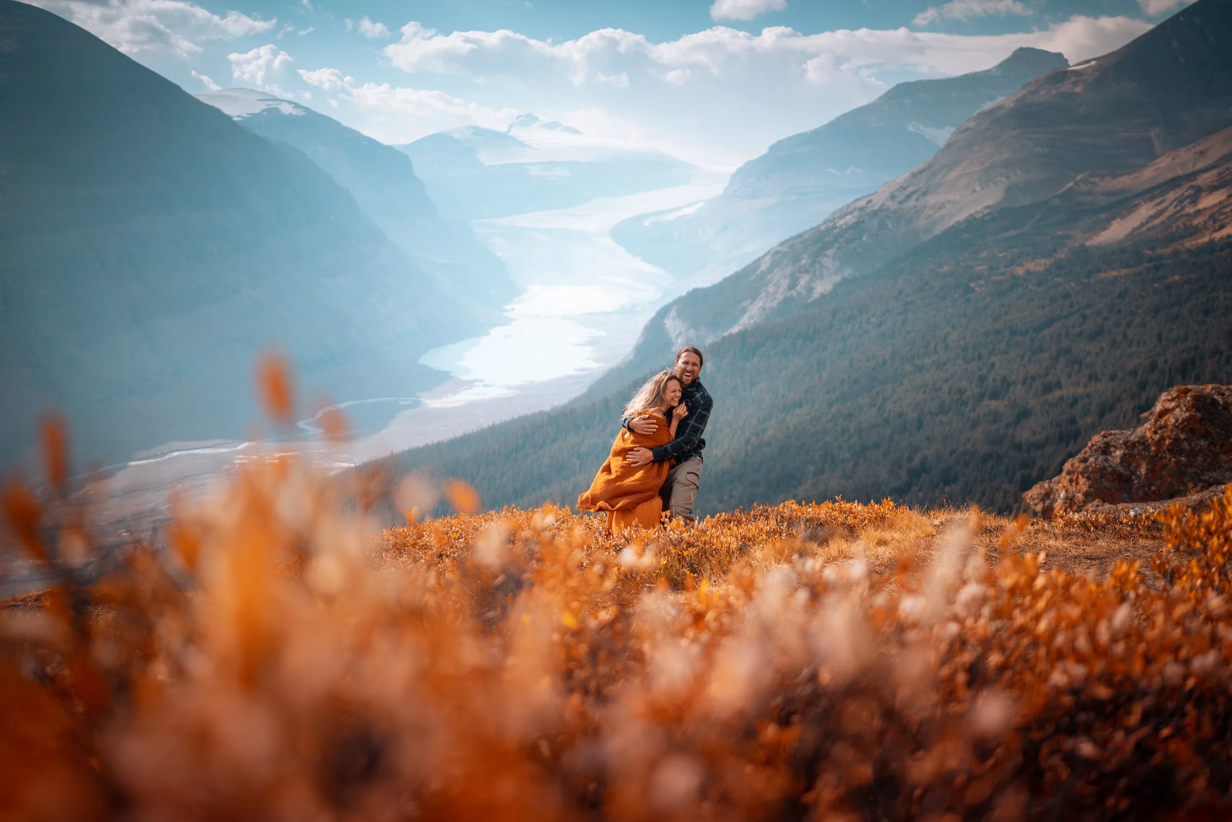 A couple hugging on a mountain in autumn, with a river and mountains in the background.