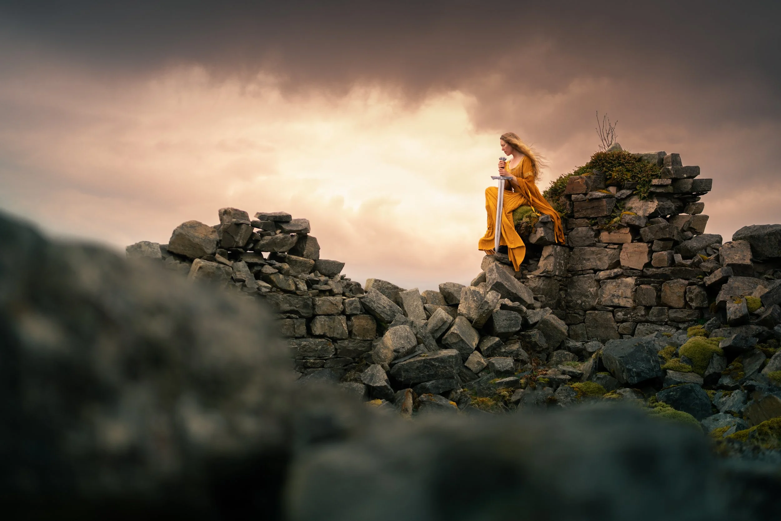 A woman in a flowing yellow dress sitting on a stone ruin holding a sword with a cloudy sky background.