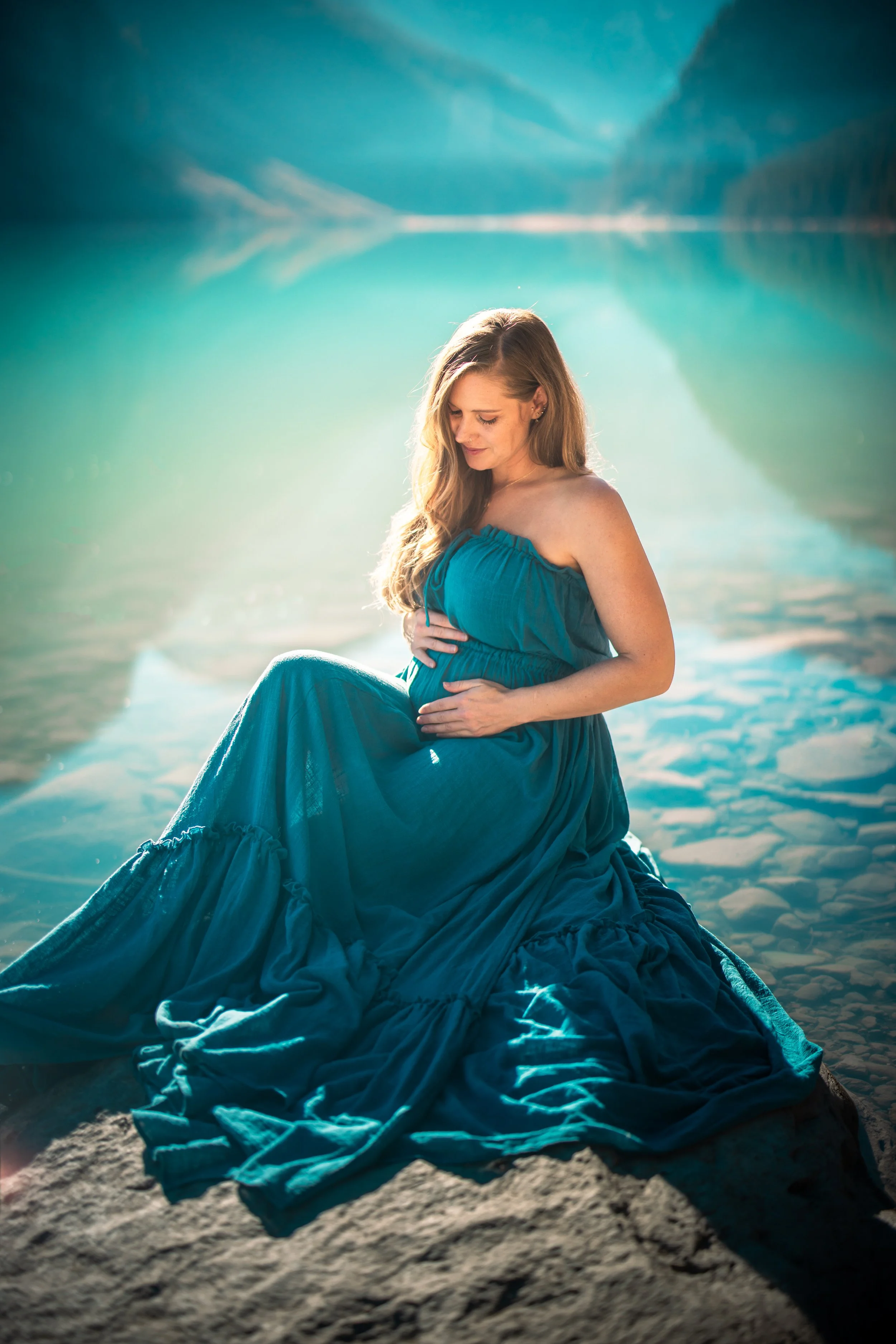 A woman wearing a long, flowing teal dress sitting on a rock by a lake.