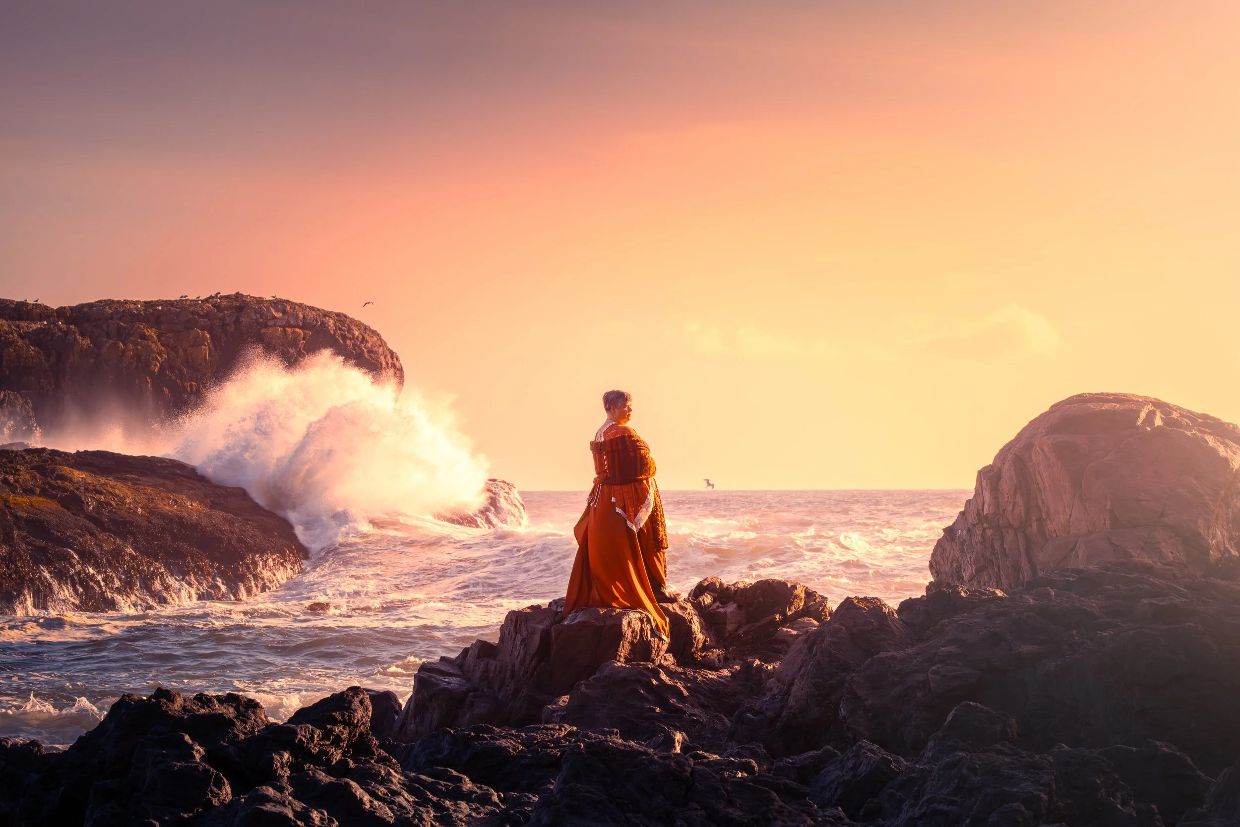 A woman in a long orange dress standing on rocks by the ocean during sunset with waves crashing against the rocks and the sky in shades of orange and pink.