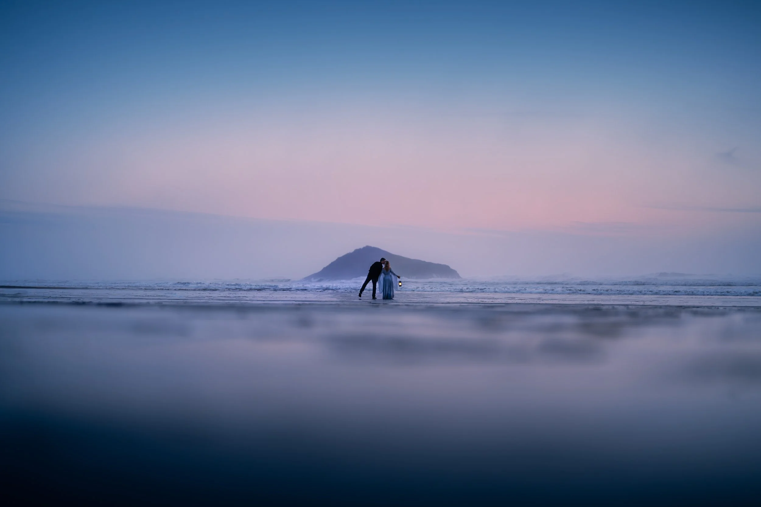 A romantic couple standing close on a beach at sunset, with an island in the background and calm ocean waves.