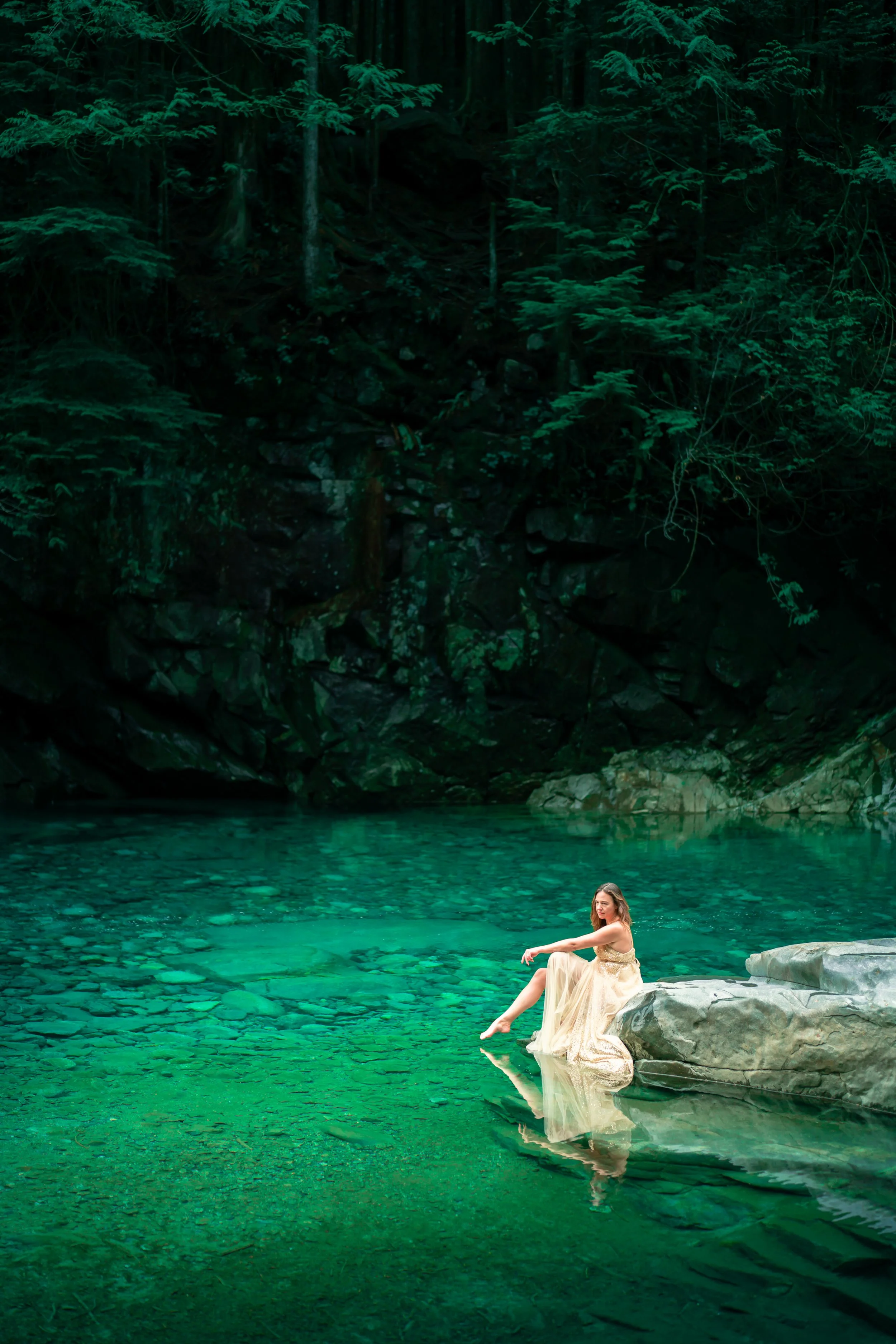 A woman in a flowing, cream-colored dress sitting on a large rock at the edge of a clear, greenish lake surrounded by a dense forest.
